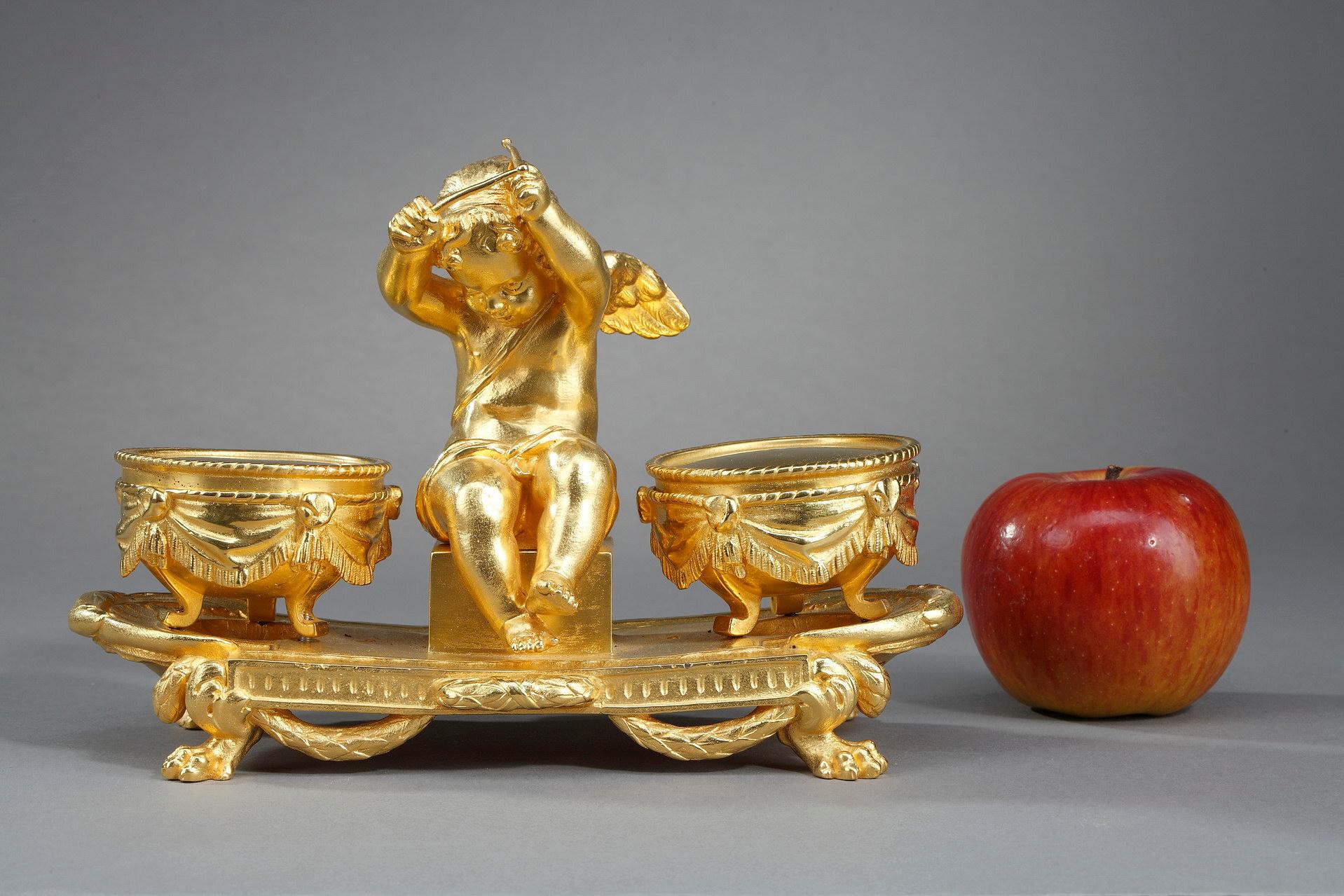 Gilt bronze inkwell in the Louis XVI style, representing Love seated with two sticks in his hands, surrounded by timbales. The circular timbales in gilt bronze, are decorated with drapery and rest on three feet. The whole is fixed on an oval base