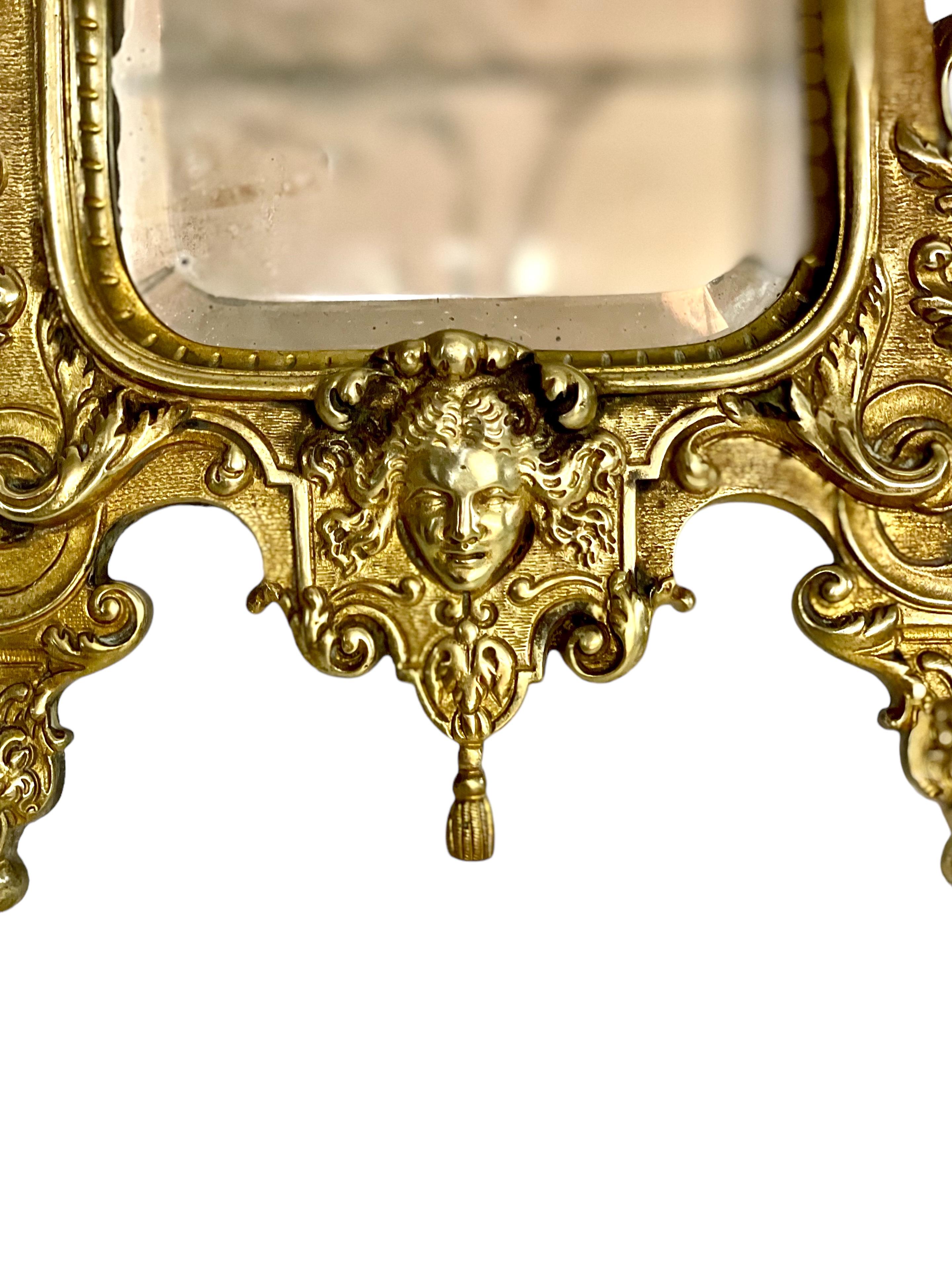 A petite but very impressive gilt bronze Rococo vanity mirror, dating from around 1900, and featuring ornately cast floral and foliage decoration around its frame. At the crest, a flower- filled urn is flanked by two flaming lamps, lapped by