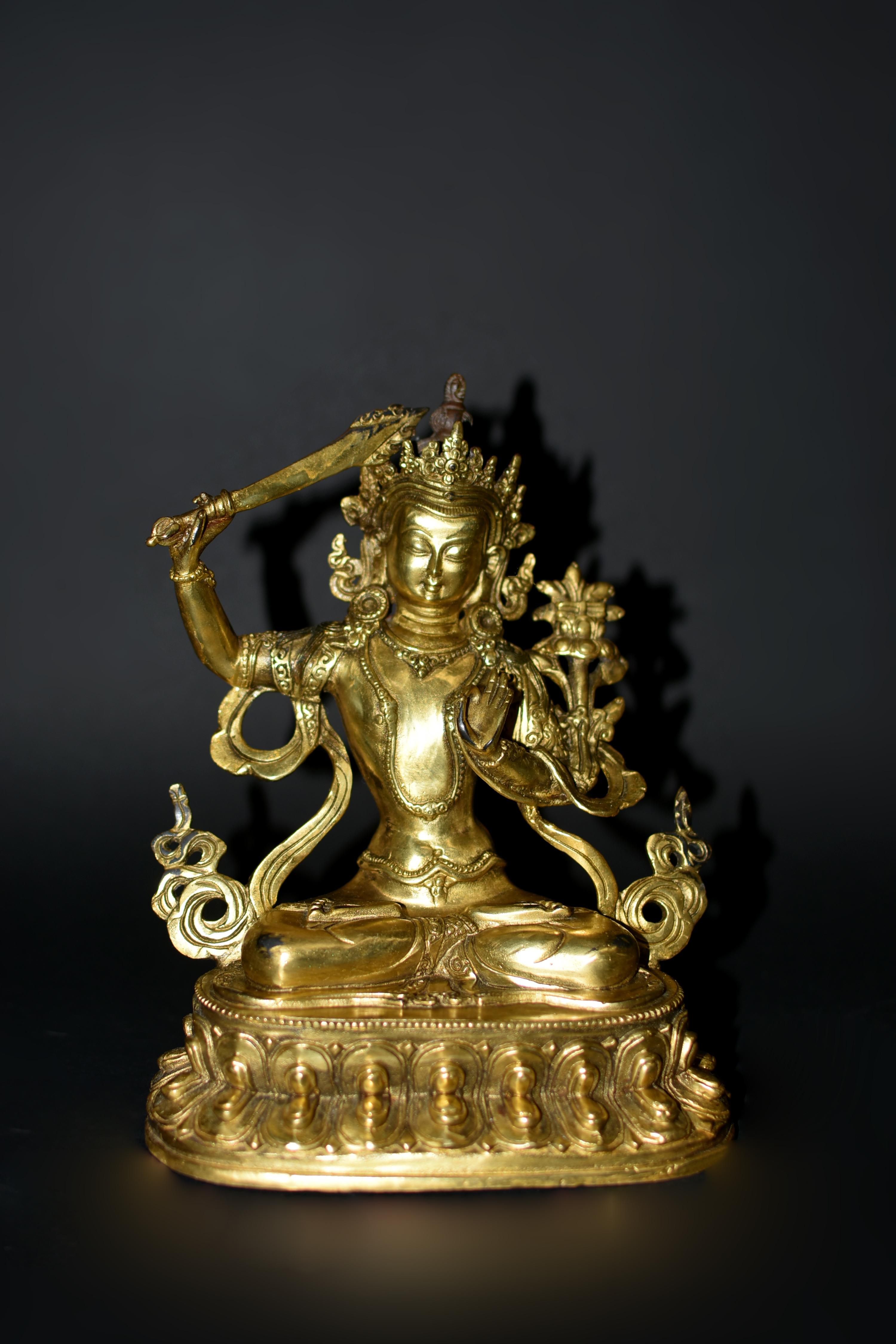 A beautiful, brightly gilded bronze statue of Tibetan Bodhisattva Manjushree. Seated in vajrasana on double lotus base, his right hand holding the sword of wisdom and his left hand in jnana mudra, gesture of teaching. The smiling face with downcast