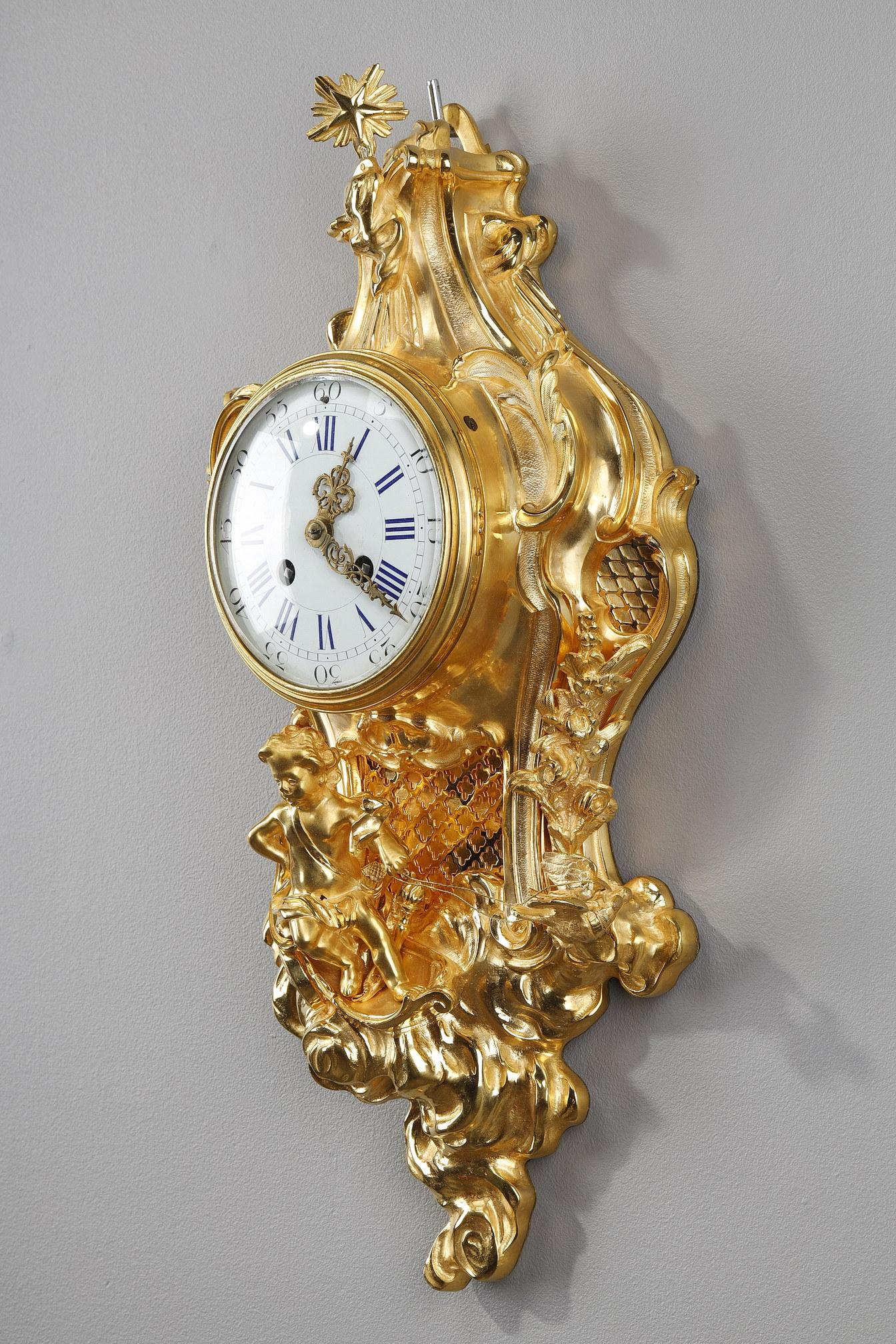 An ormolu alcove cartel in the Louis XV style presenting a Love on a chariot pulled by a couple of doves in the clouds on a background of trellises. The rocaille decoration is topped by a star. The dial is enamelled in white with Roman numerals for