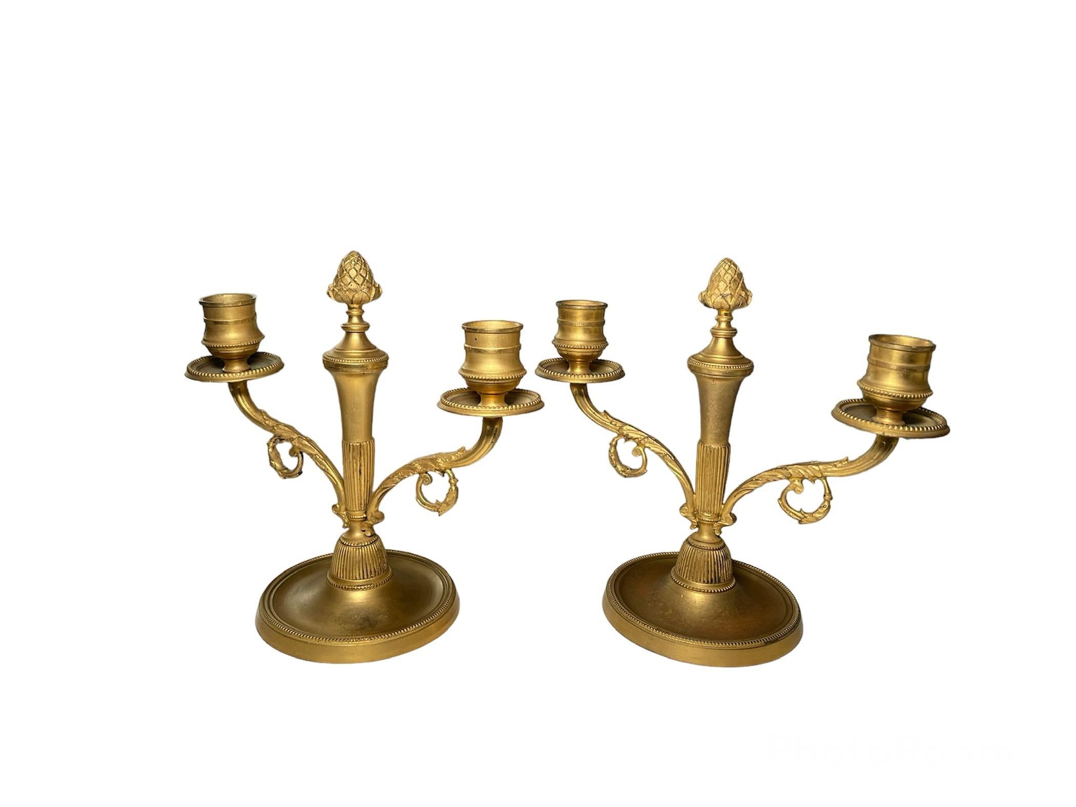 This is a pair of gilt bronzed metal double candle holders. They depict double candle holders decorated with some scrolls of acanthus leaves in their branches and pine cones in the center of them serve as a finial. They stand over round bronze metal