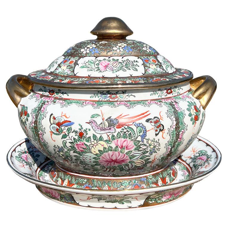 A large oval chinoiserie ceramic tureen with lid and underplate. This ceramic dish is hand-painted and decorated with a grade scene. On each side, a brightly colored bird perches upon a bouquet of pink, yellow, green, and blue flowers. Each side of
