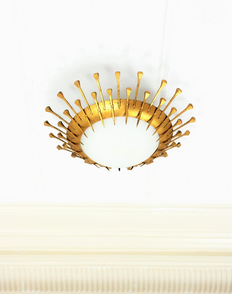 Spectacular hand-hammered iron and opaline glass sunburst flush mount / wall light. France, 1940s-1950s.
This sunburst light fixture features alternating short and long nail decoration and gold leaf finish. Style in transition from Art Deco to