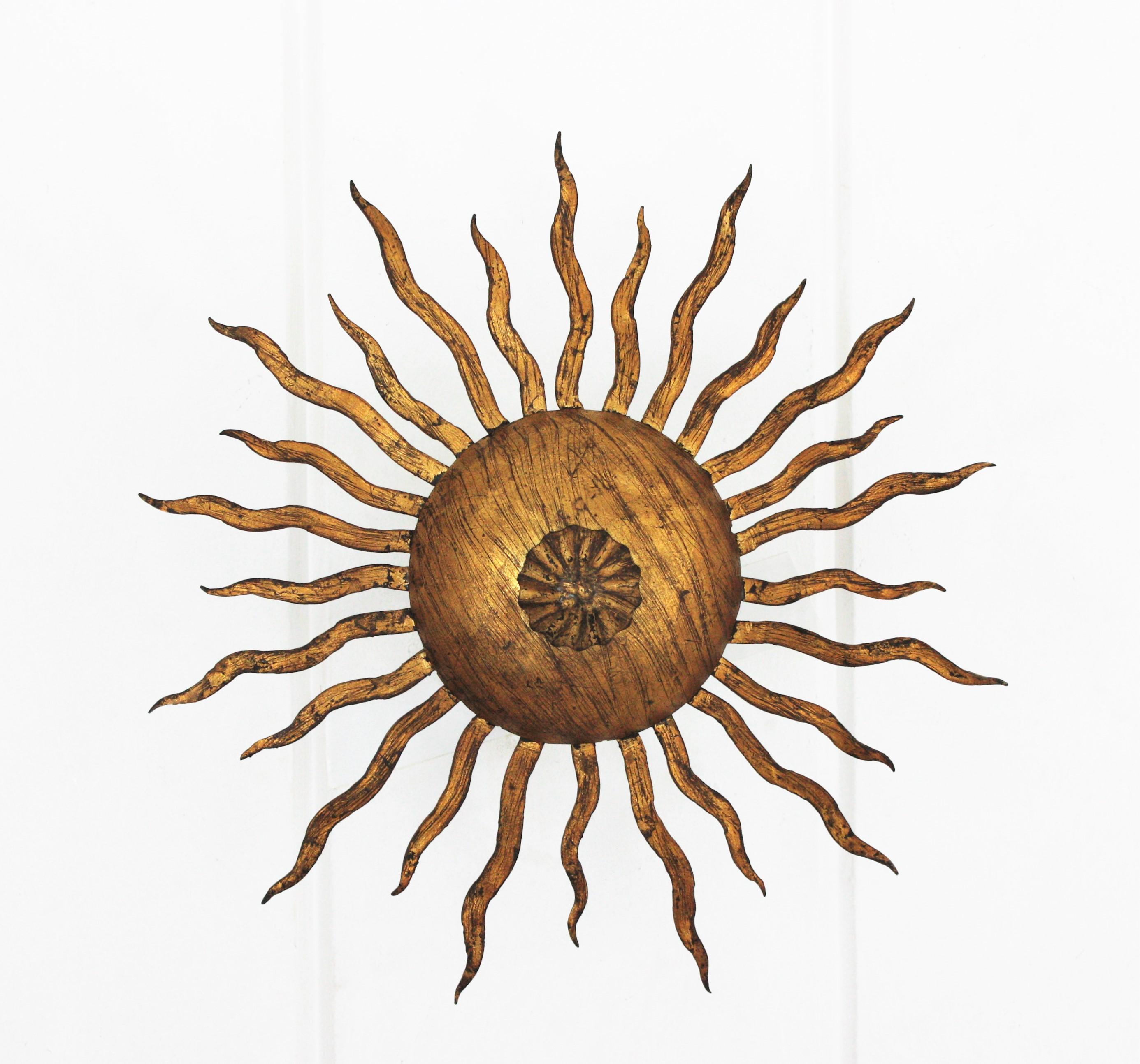Wrought gilt hand forged iron sunburst flushmount ceiling lamp or light fixture. France, 1950s.
This hand-hammered iron flushmount has alternating rays in two sizes surrounding the central sphere and a flower decoration at the central part.
This