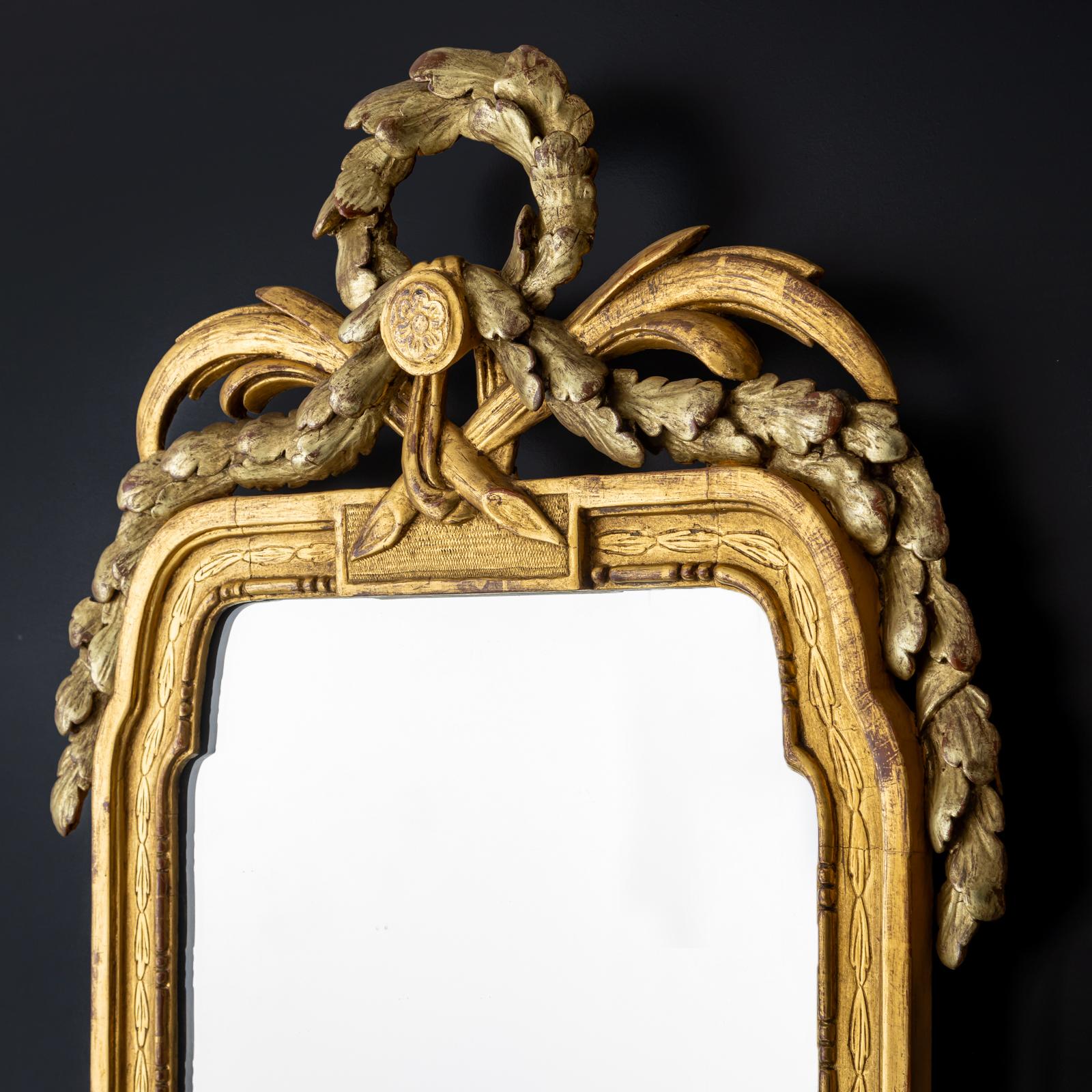 Miroir mural doré en bois sculpté avec applications de stuc. Le cadre rectangulaire haut est arrondi au sommet et couronné d'un feston de couronnes de laurier en argent patiné. Les mêmes festons complètent la section inférieure qui s'incurve vers