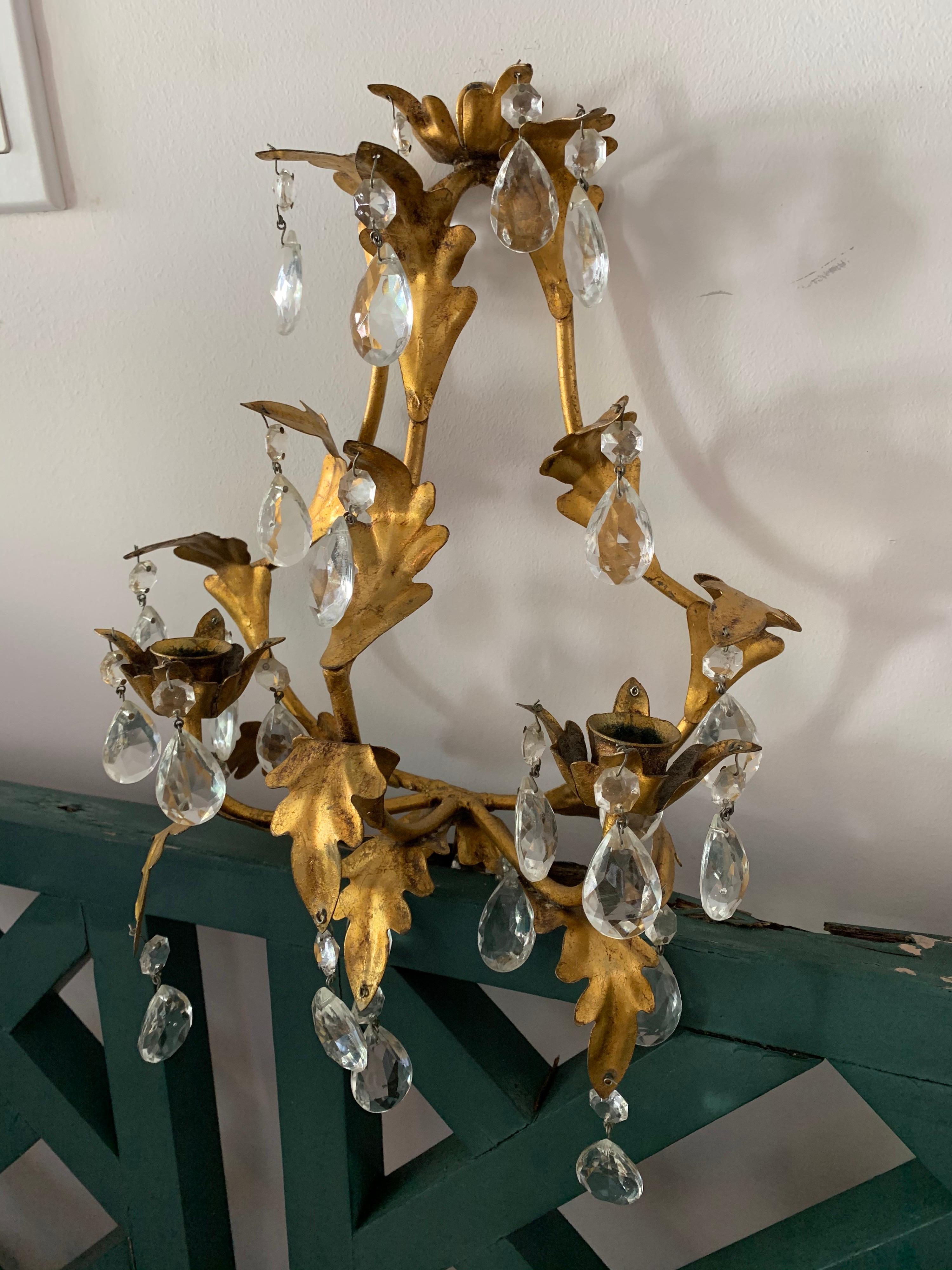 A single gilt metal wall mount candelabra sconce with two arms and glass pendant drops.

Designed to hold two candlesticks. Not electrified.

Dimensions: 11 inches W x 6.5 inches D x 15 inches H.
 