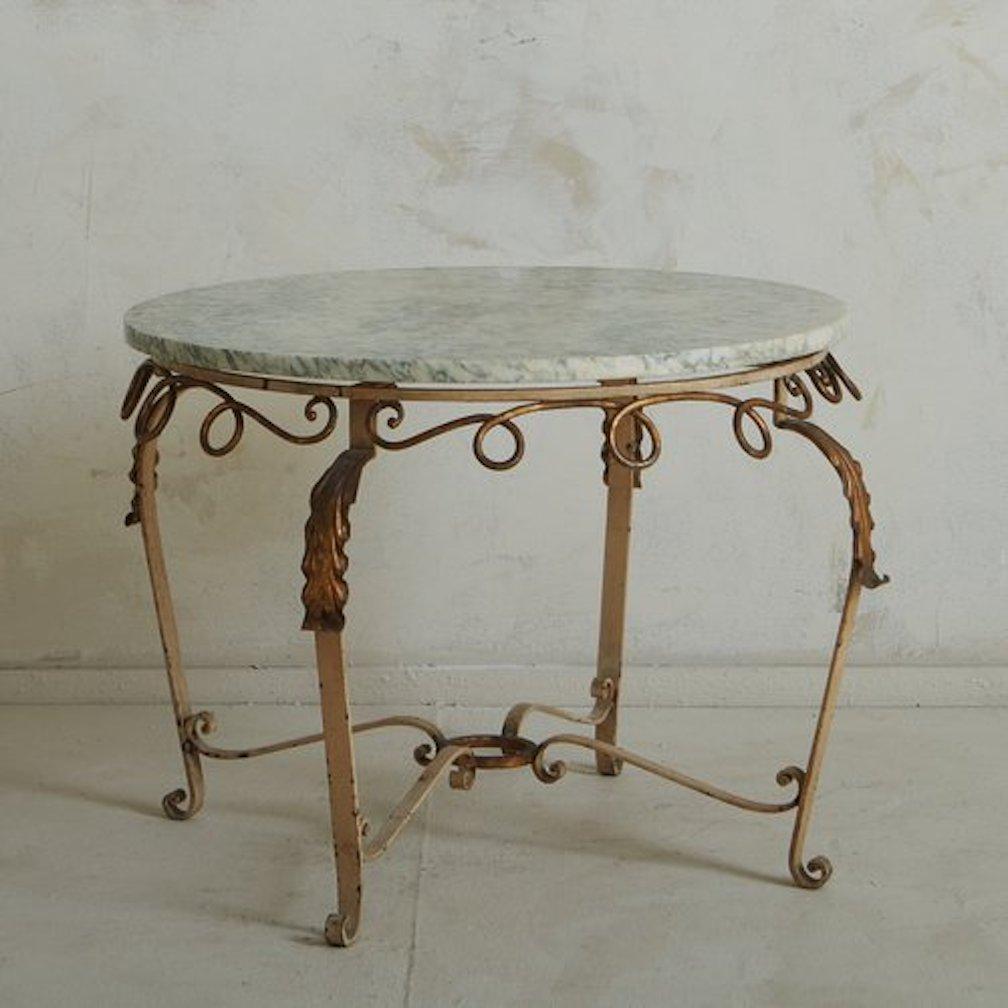 A 1970s French coffee table featuring a cream painted metal base with four curved legs and scroll stretchers. This table has scroll detailing and overlaid leaf motifs at the top of each leg, which have a gold gilt finish. It has a circular cream