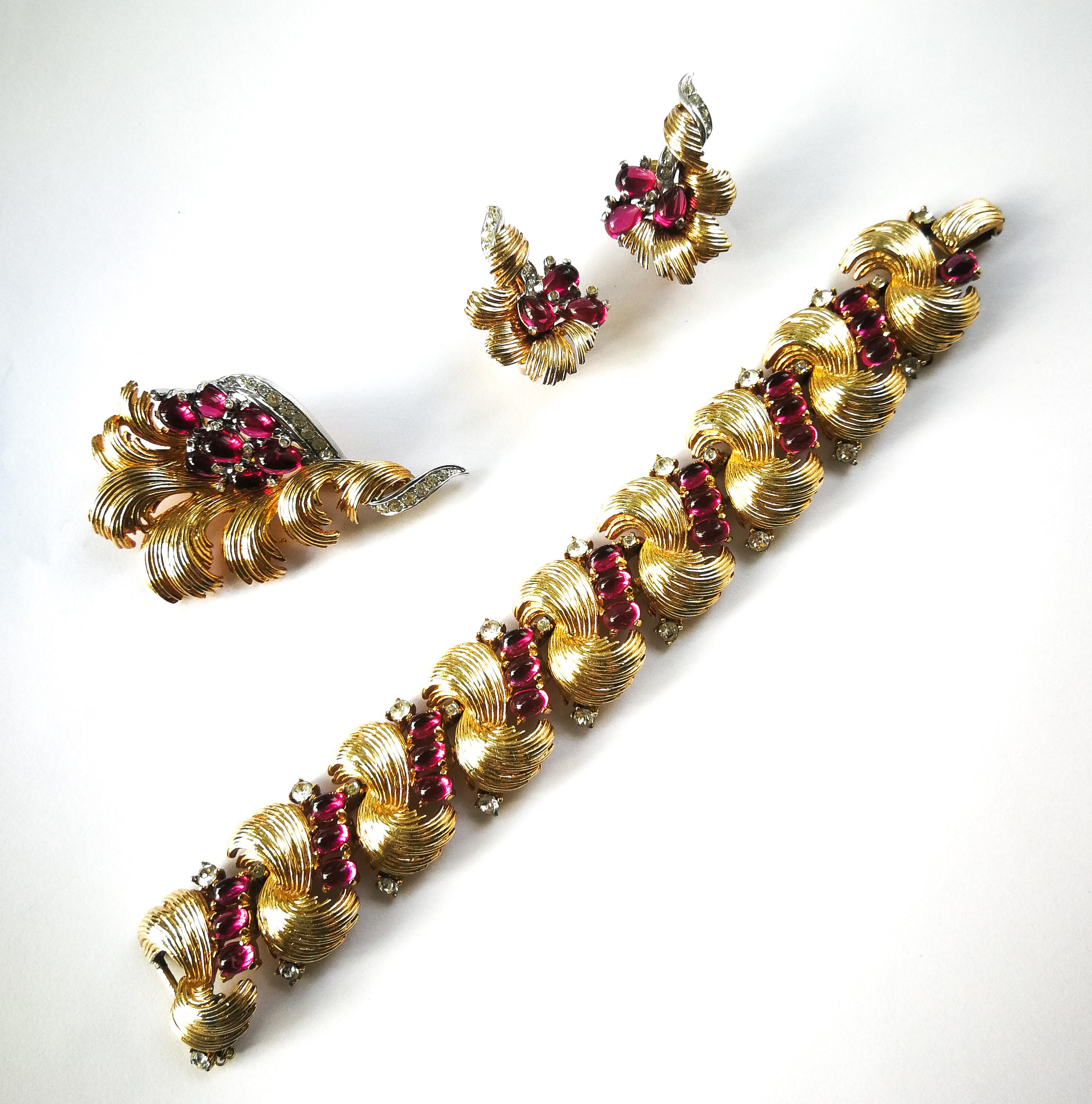 An exquisite and sumptuous three piece set by Marcel Boucher, from the 1960s. An articulated bracelet, with safety chain, a brooch and earrings make up this parure, in an exceptional design, with ruby glass cabuchons set in gilded feather like