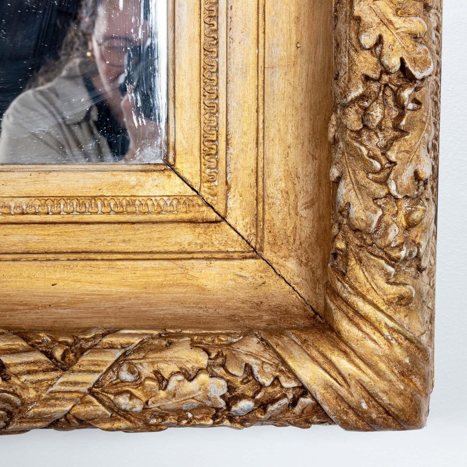 A generously sized English mirror with a gilt gesso frame. Crafted in the early 20th century, this mirror bears the hallmark of English craftsmanship at its finest. The frame, adorned with a repeating oak leaf pattern, dances with elegance and