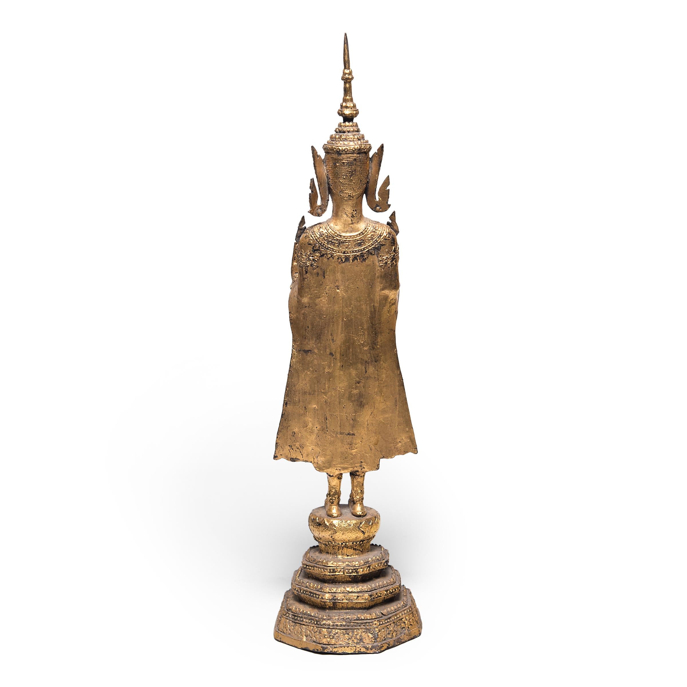Dated to the mid-19th century during the Rattanakosin dynasty, this serene standing Buddha figure is intricately cast in bronze and decorated with gold leaf. The standing figure is costumed in bejeweled robes reminiscent of Thai royal attire and