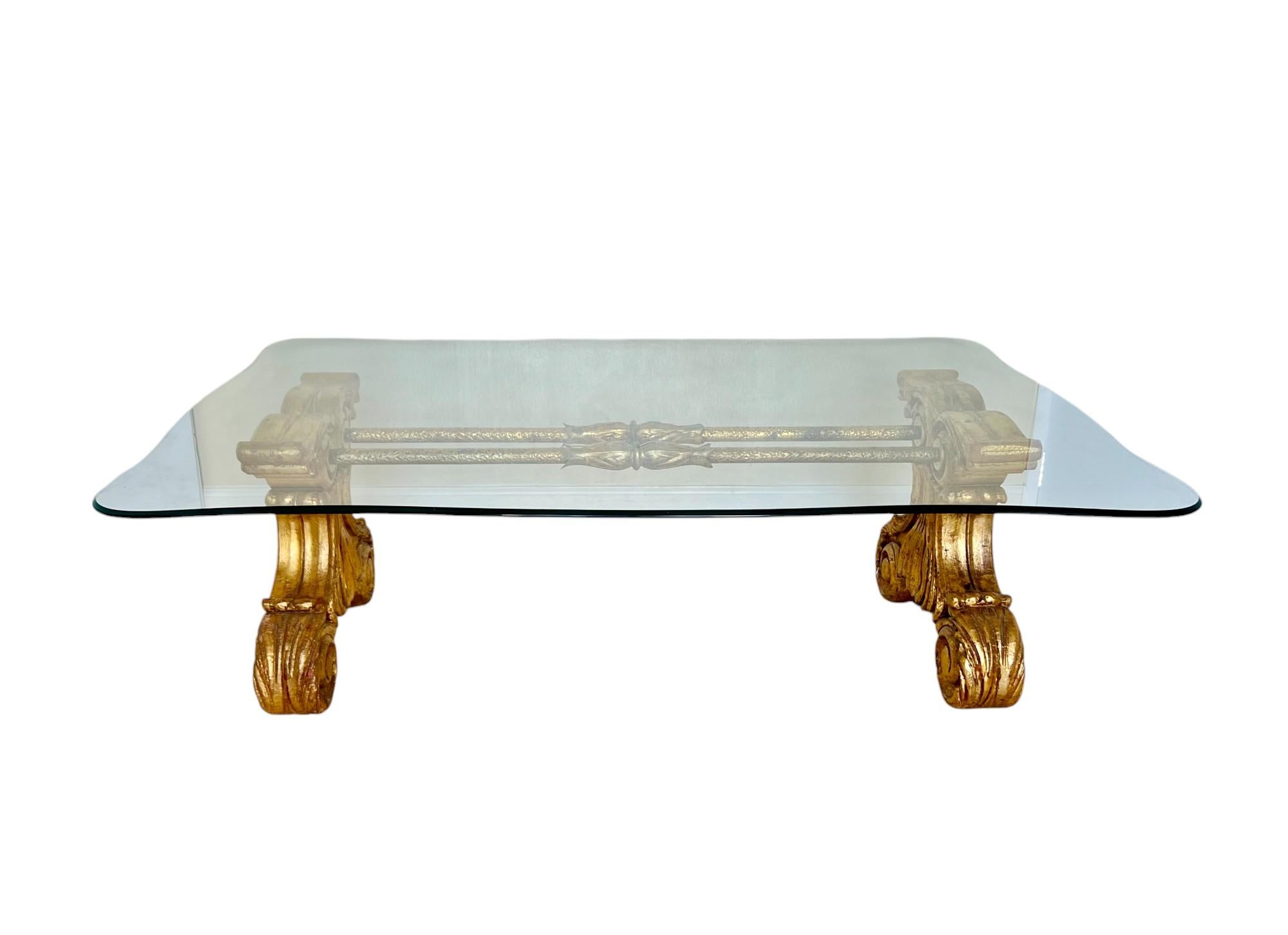 Table basse ancienne en bois doré et métal avec plateau en verre. Cette magnifique pièce présente des extrémités en bois sculpté doré avec un motif de volutes, reliées par deux tiges en métal doré avec des détails martelés. Plaque de verre de forme