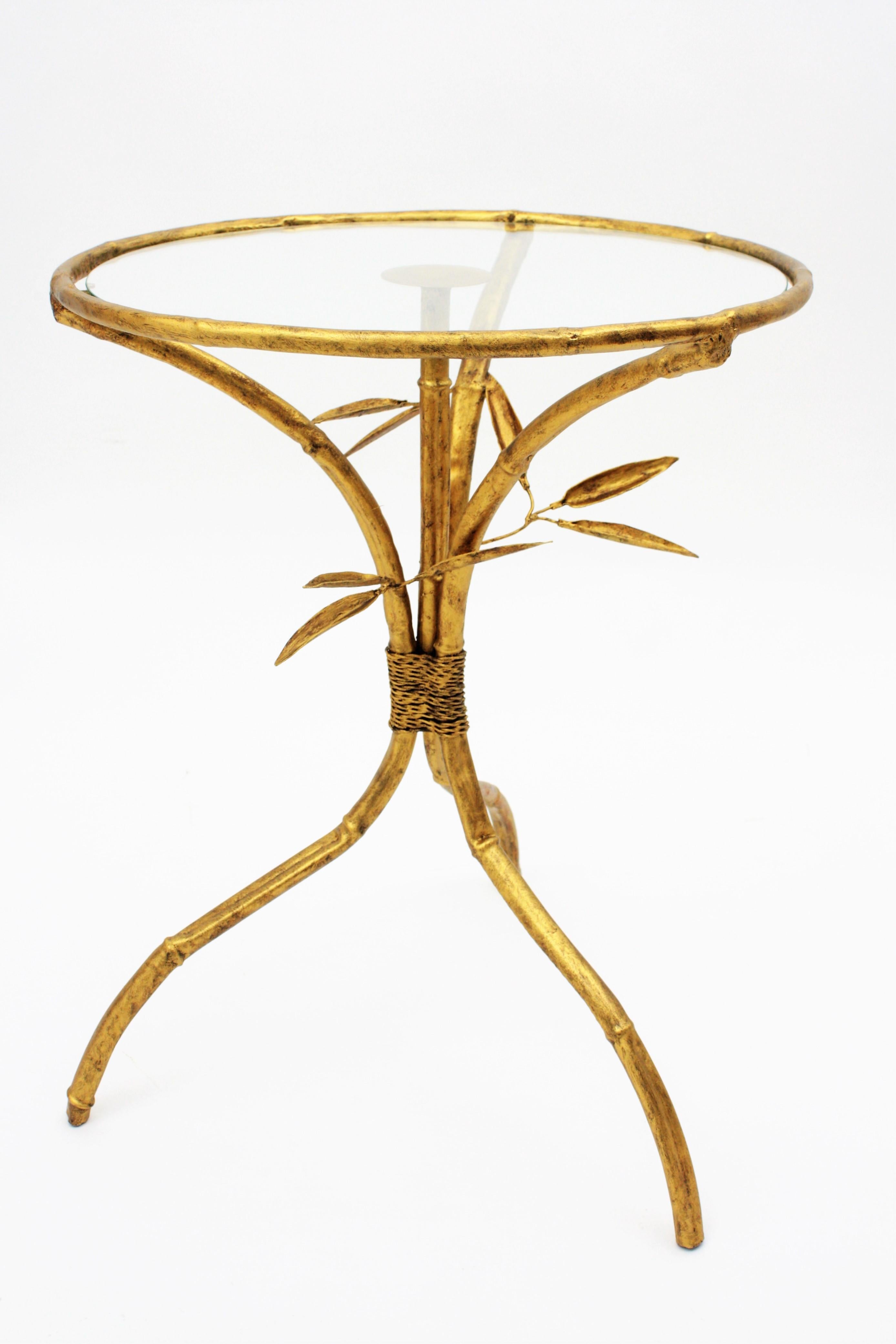 Elegant Hollywood Regency gilt iron faux bamboo gueridon drinks table with round glass top and tripod base. Spain, 1950s. 
This table is made in hand-hammered iron gold leaf finish. It has a beautiful faux bamboo design with leaf and rope details
