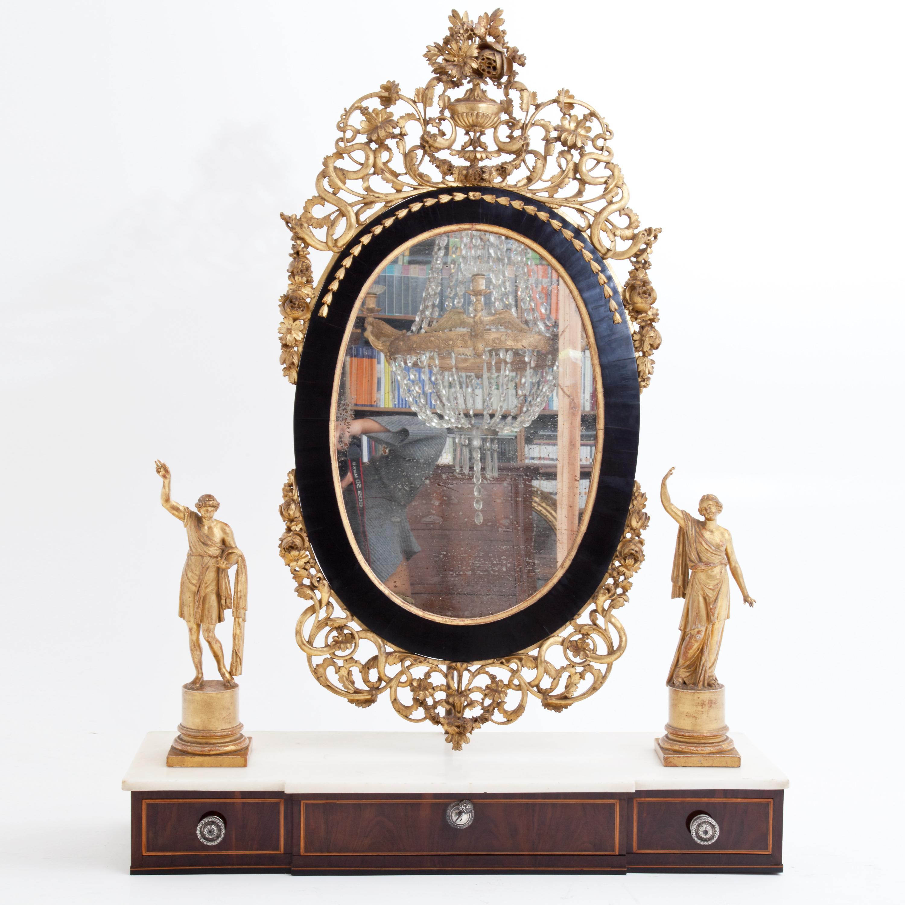 Mirror mounted on a wide drawer with silver handles in an oval black frame and rich tendril and flower decoration made of giltwood. The mirror is flanked by figurative bronzes in antique habit mounted on the marble top. The drawer front is veneered
