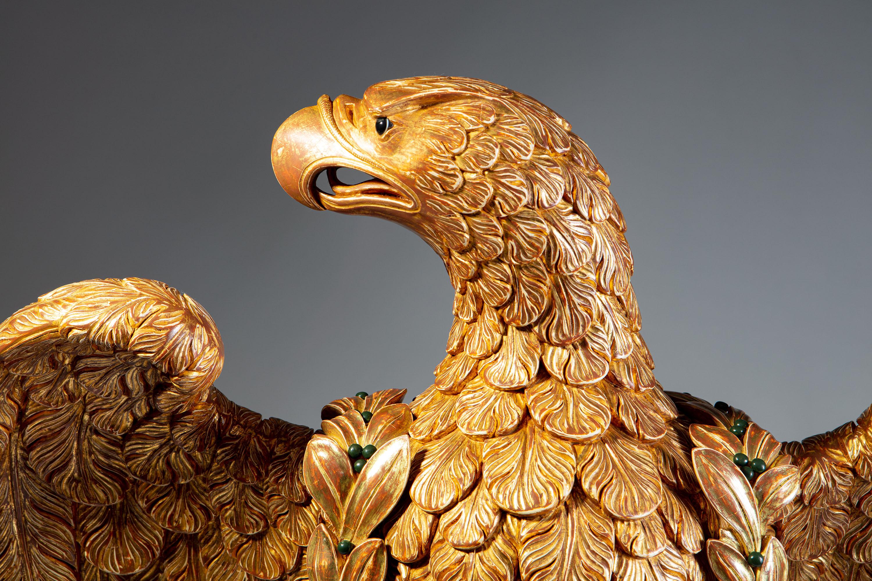 Ein prächtiger und reich geschnitzter Adler aus Goldholz, der mit geöffneten Flügeln auf einem Felsen sitzt. Der Kopf des Adlers blickt nach links und ist mit einem separaten Kranz aus Goldholz versehen, der mit Cabochons aus Malachit und Achat