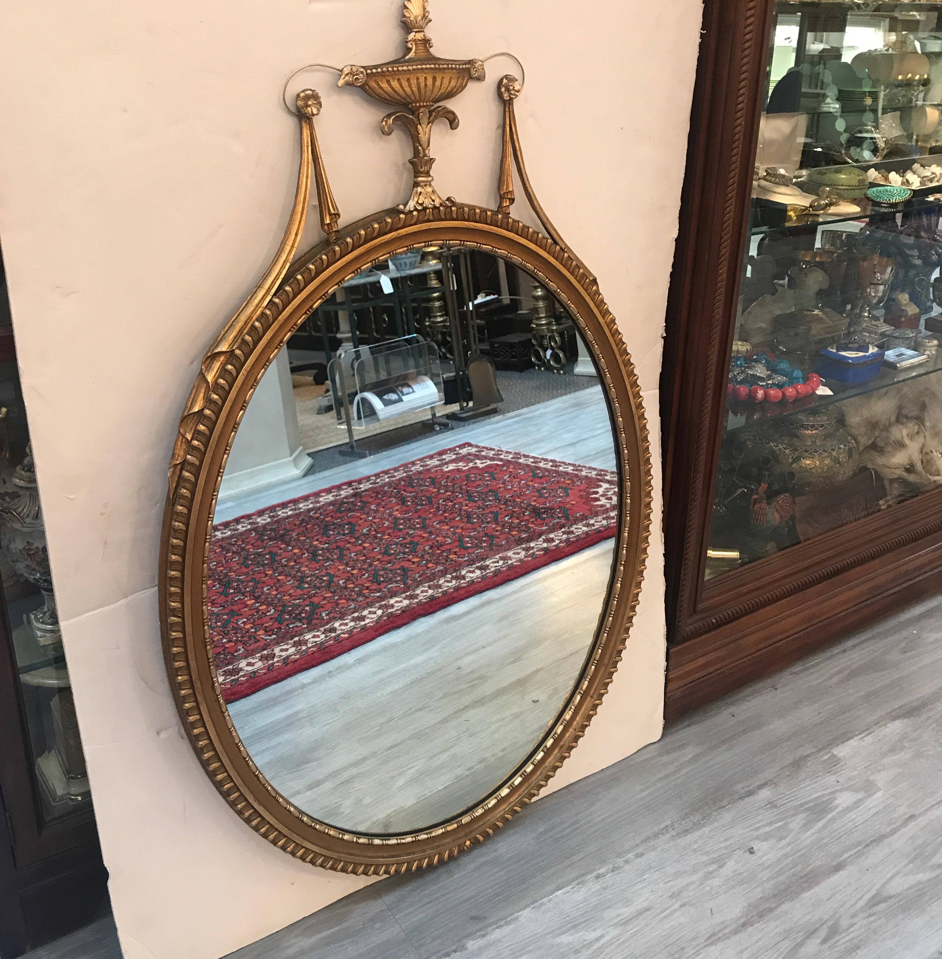 A graceful oval Adam style giltwood mirror with urn and swag pediment top. The frame with gadrooned edge all around. The gold in excellent condition with some age patination, the silvering on the mirror is excellent. The mirror alone measures 30