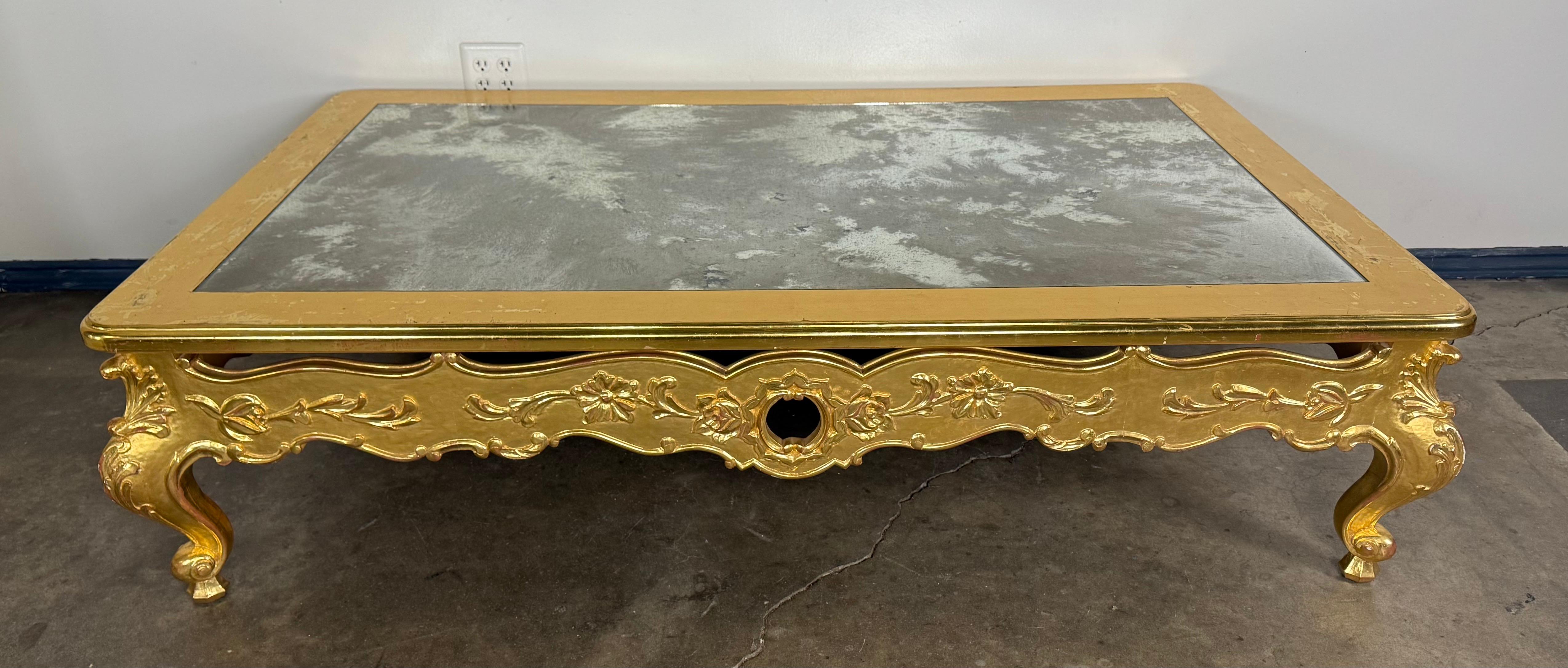 A striking Rococo-style giltwood coffee table featuring an inset antiqued mirrored top framed by a molded gilt edge. The apron is richly carved with scrolling foliate and floral motifs, centering on a cartouche design and raised on elegant cabriole