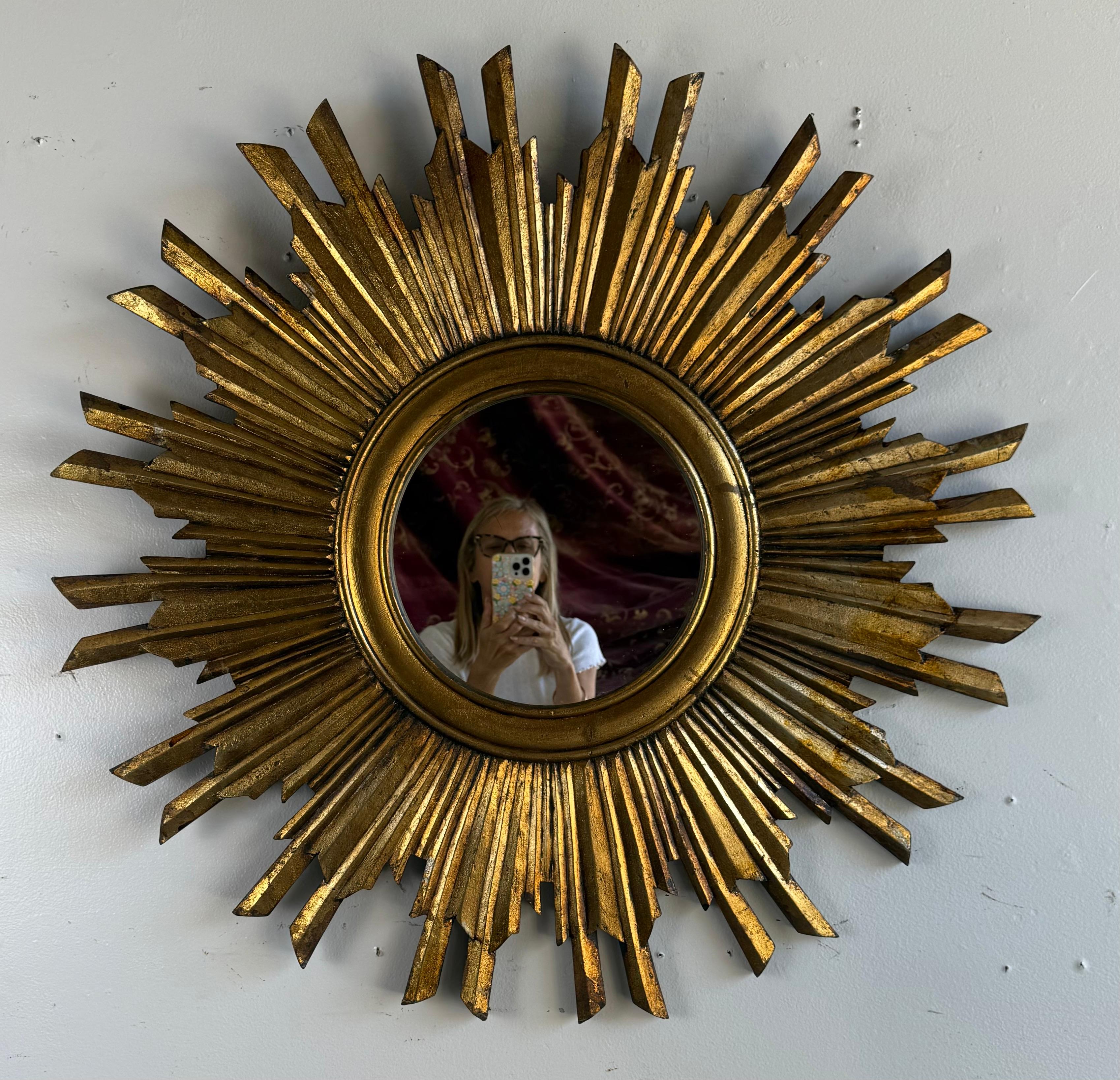 A striking sunburst mirror, mid-20th century, featuring a bold radiating frame of carved wooden rays in varying lengths and angles, emanating from a circular mirror plate. The surface is finished in a rich hand-applied gold leaf with natural age,