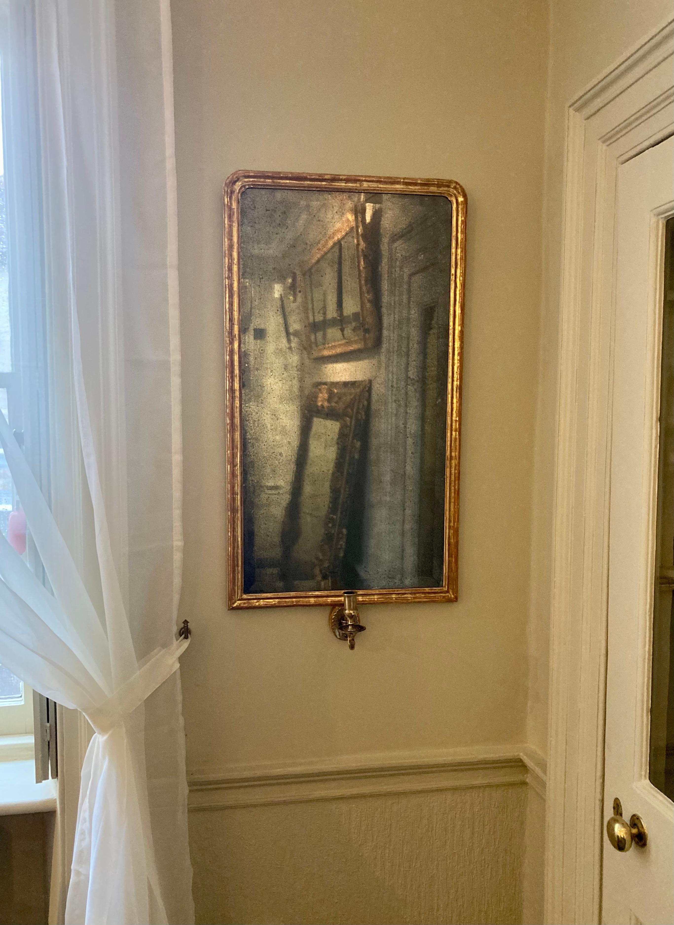 A highly decorative  rectangular giltwood wall mirror with an 18th Century bevelled mercury glass with gentle foxing throughout surrounded by a later giltwood rectangular frame with softened round edges to the top and single brass candle arm to the