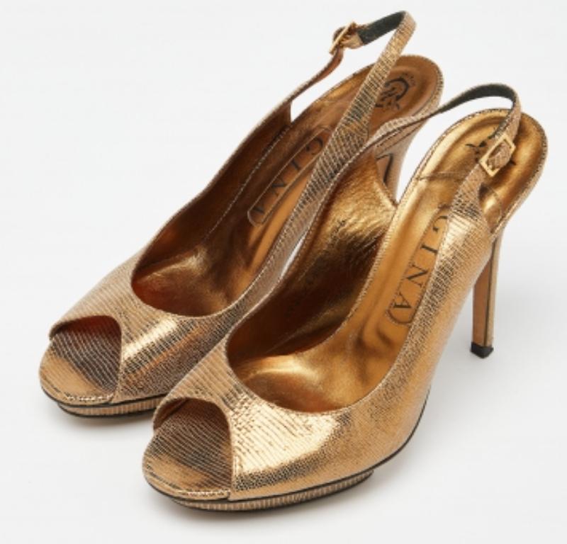 Questi deliziosi sandali Gina sono ideali sia con i vestiti formali che con quelli casual. Realizzate in pelle lucertolata color oro, si abbinano a punte di elefante, tacchi di 12 cm e slingback. Sono rifinite con solette e plateau foderati in pelle