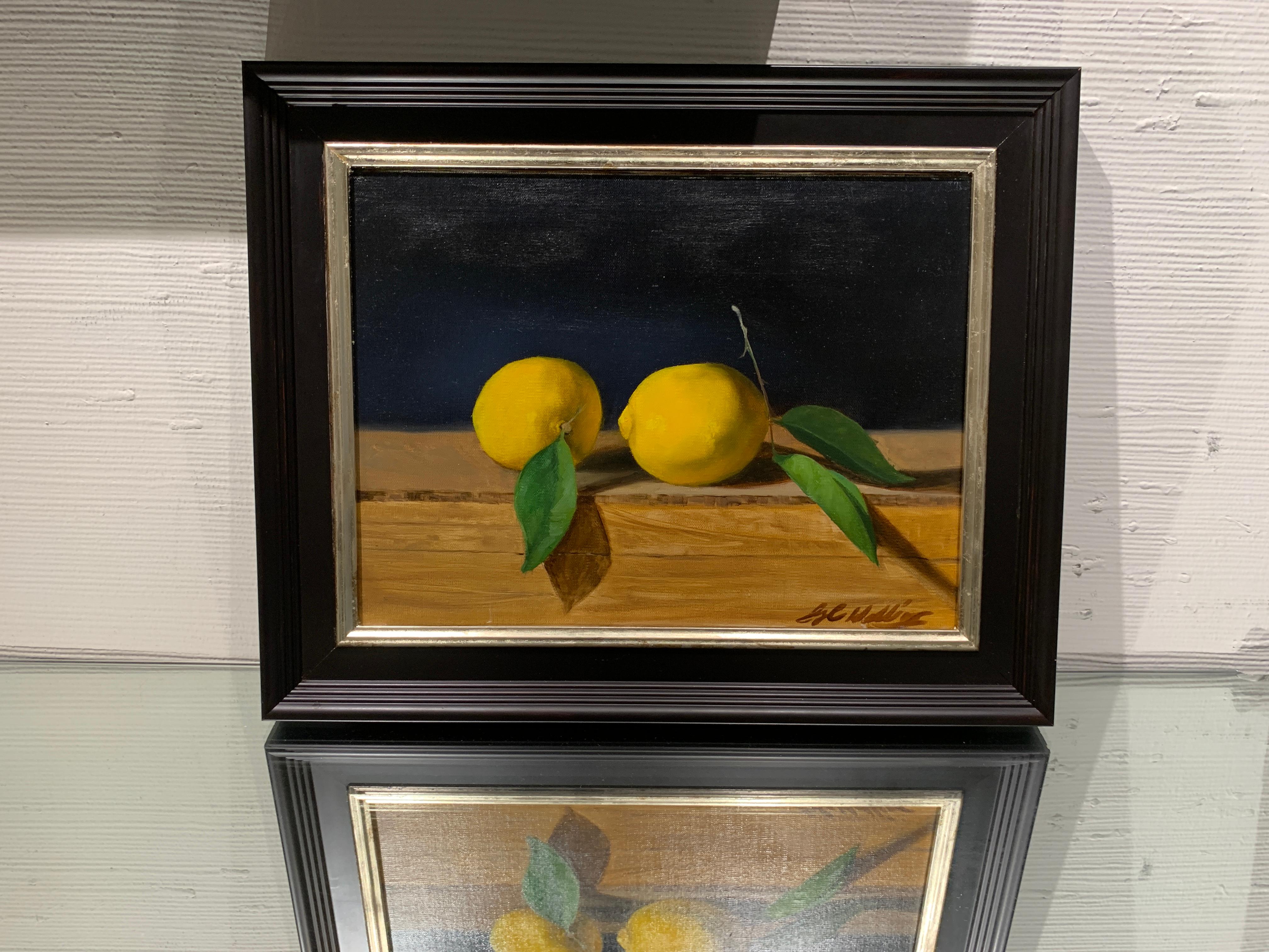This realist still life painting by American artist Ginny Williams is the depiction of two lemons laying on a wooden table with a rich, black background.  This piece is framed in a traditional black frame with a silver fillet.  The unframed size is