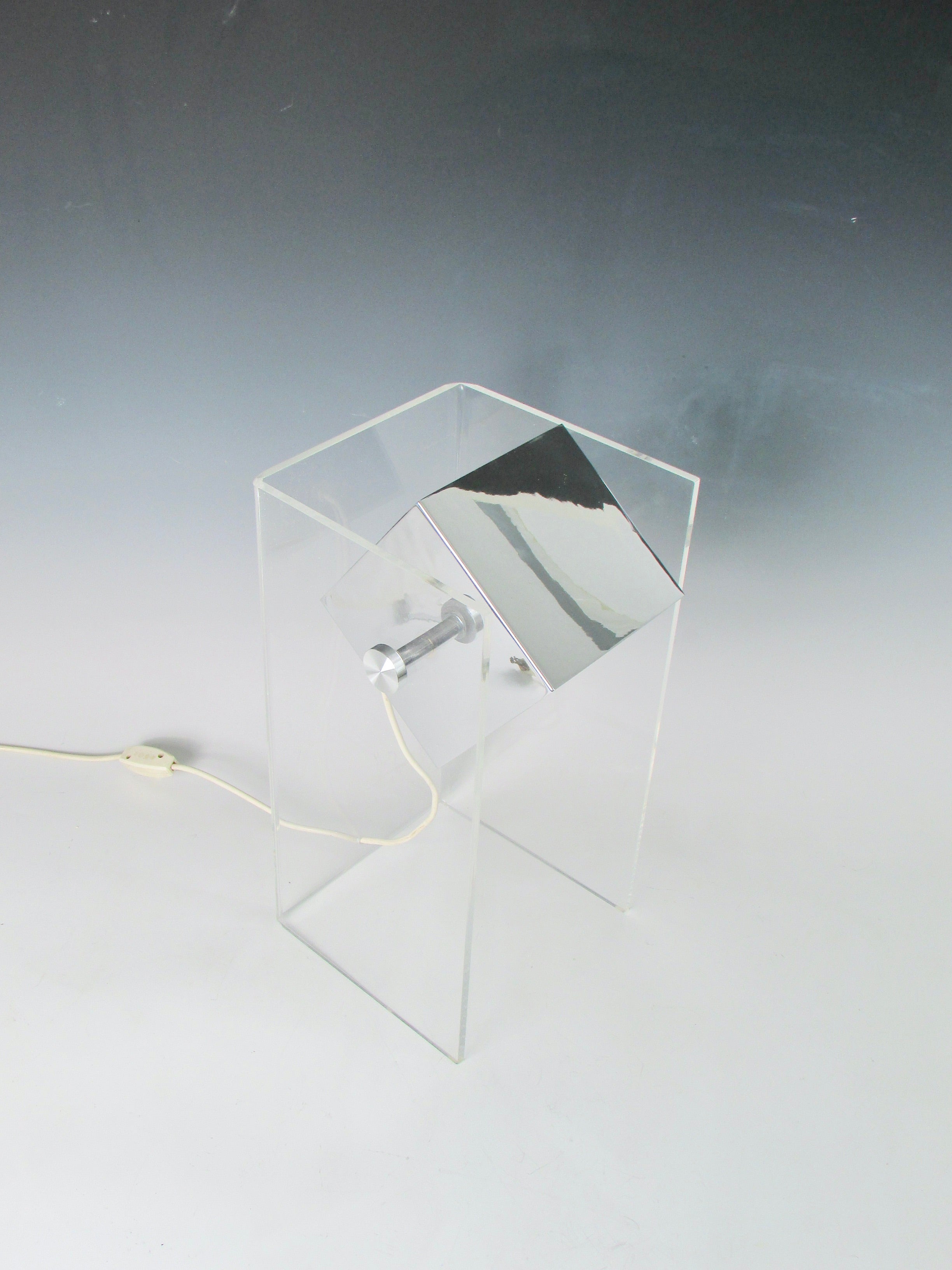 Simply designed square chrome  housing set  in rectangle acrylic base table lamp. Up ended Lucite rectangle holds polished chrome lamp box. Lamp does tilt up and down. Chrome cube measures 5.25 square.
