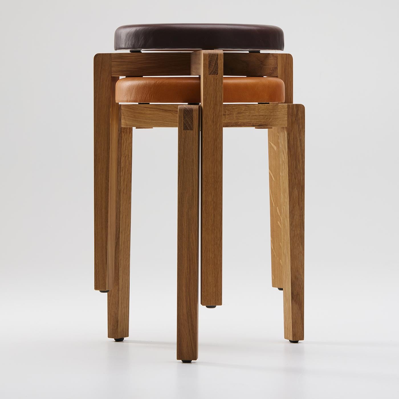 The gio stool features a round seat upholstered in cognac-colored leather, paired with a solid oak wood frame. Designed by Enrico Tonucci, it measures 30 cm in diameter and 42 cm in height, combining a compact footprint with durability. Its