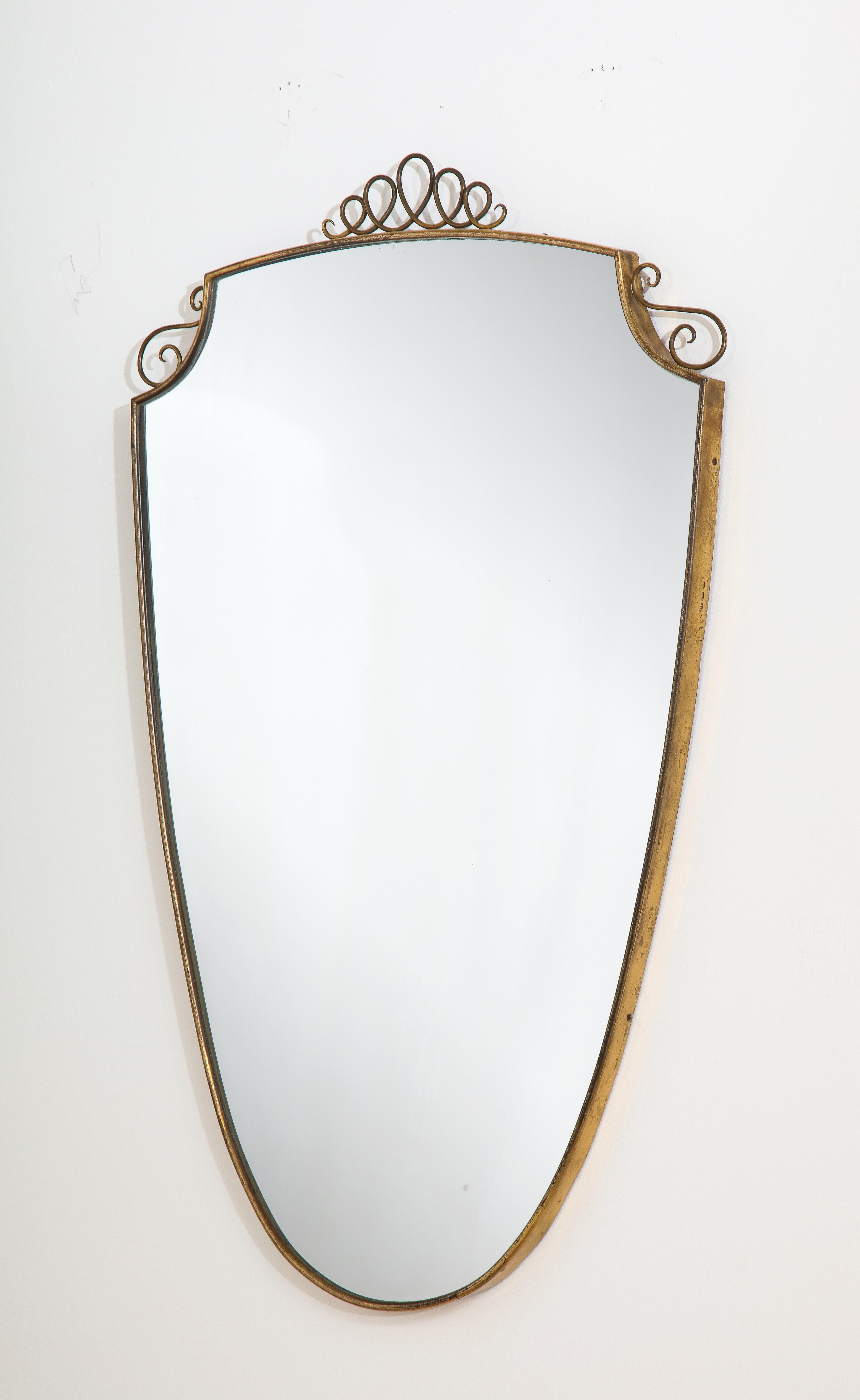 An elegant Gio Ponti brass shield shaped mirror with curly decorative motif surmounted on the crest and on each side. Ponti designed similar mirrors with the same decorative brass wave motif, in 1950, for the Hotel Bristol in Murano. With its