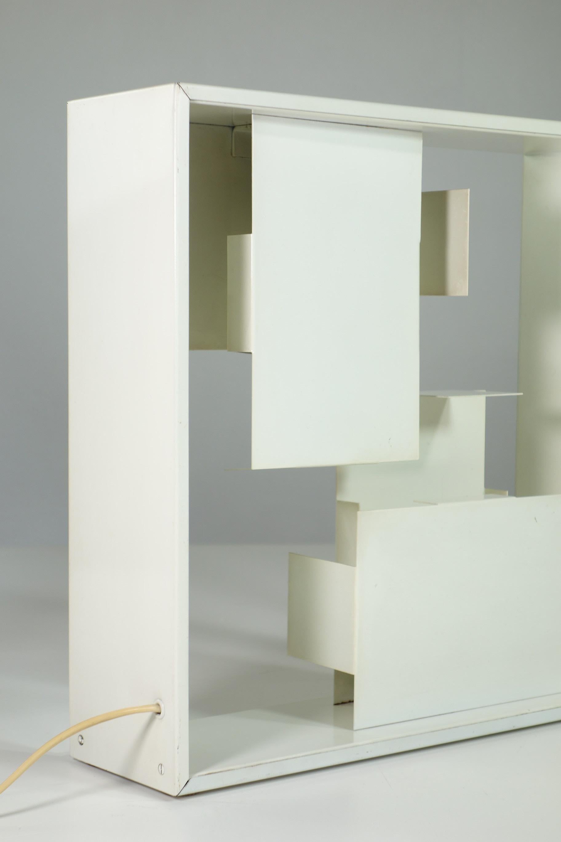 White square frame with folded geometric metal forms creating light and dark

Iconic light object and a beautiful lamp.
First production Fato lamp designed by Gio Ponti and produced by Artemide in 1967. 
A distinct characteristic lies in the