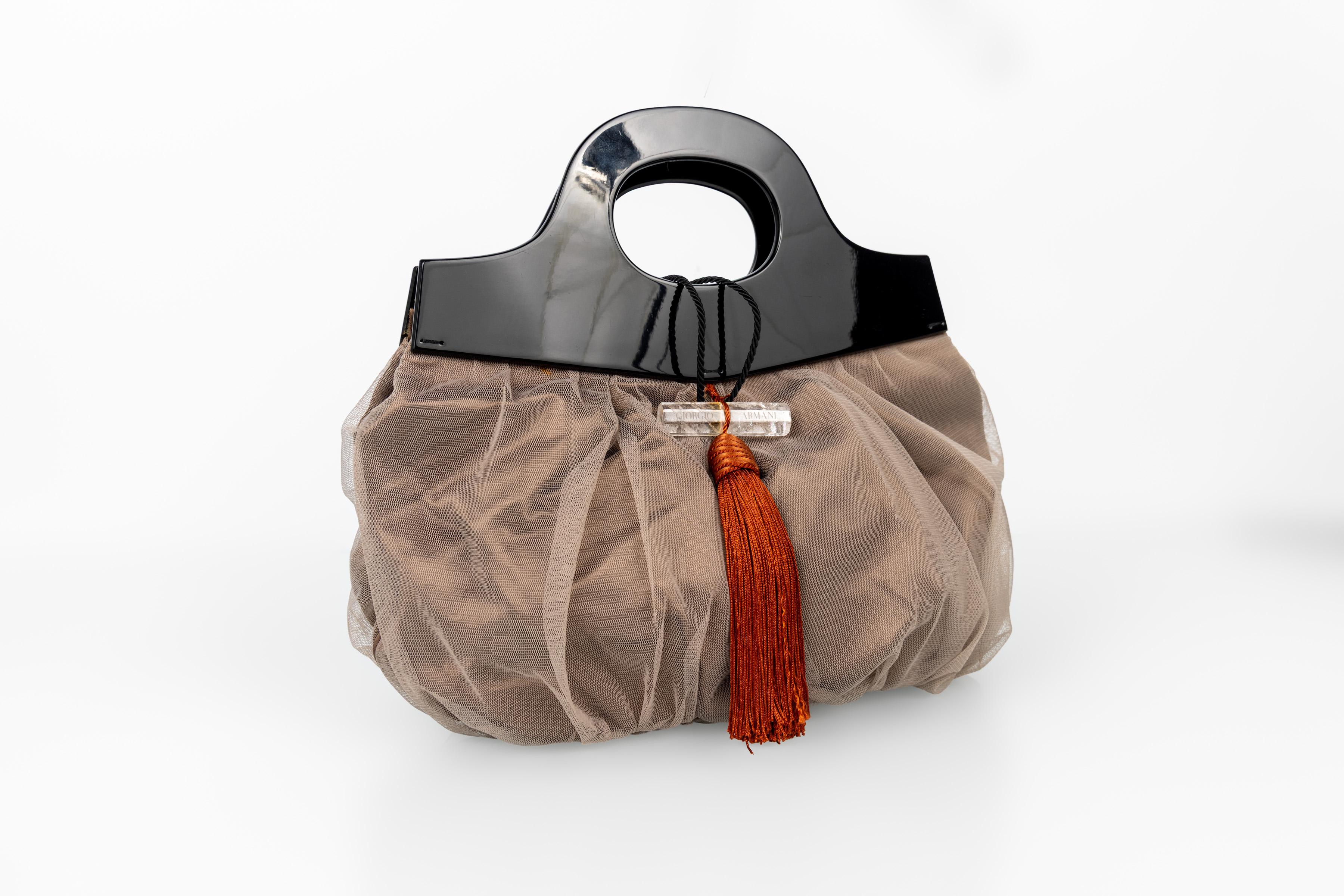 Un sac du soir raffiné et sculptural de Giorgio Armani, qui incarne le luxe tranquille et le minimalisme architectural de la maison. Le modèle se compose d'une base en soie taupe légèrement froncée et enveloppée de tulle transparent, créant ainsi