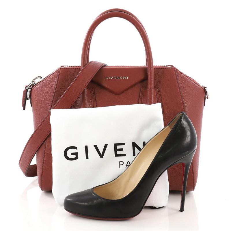 This Givenchy Antigona Bag Leather Small, crafted from brick red leather, features the signature envelope fold logo in front, dual top handles, and silver-tone hardware. Its top zip closure opens to a beige fabric interior with side zip and slip