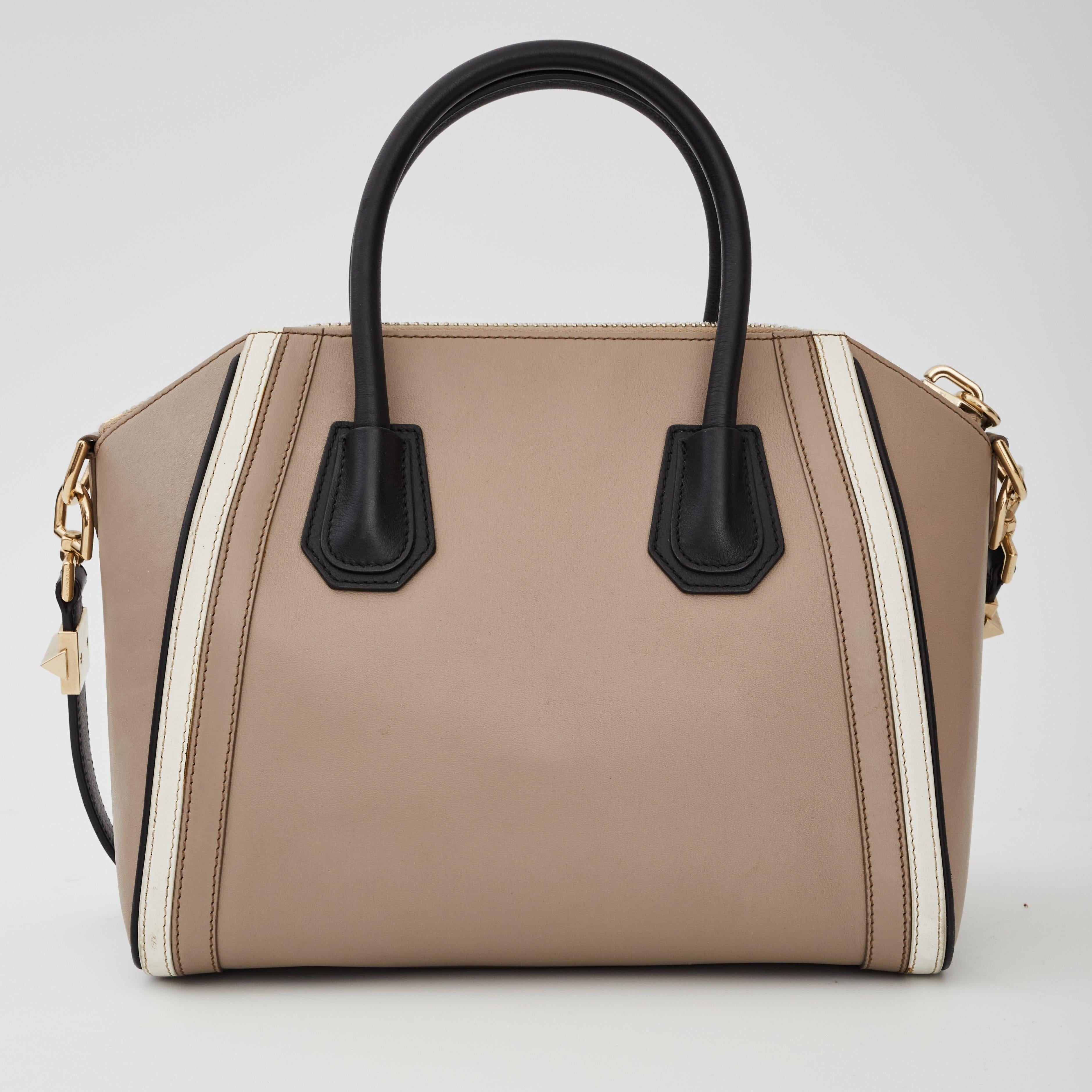 This tote is made of luxurious smooth taupe calfskin leather with white and black leather trim. The bag features thin and sturdy rolled leather leather top handles, an optional shoulder strap, gold hardware and expansive sides. The top opens with a