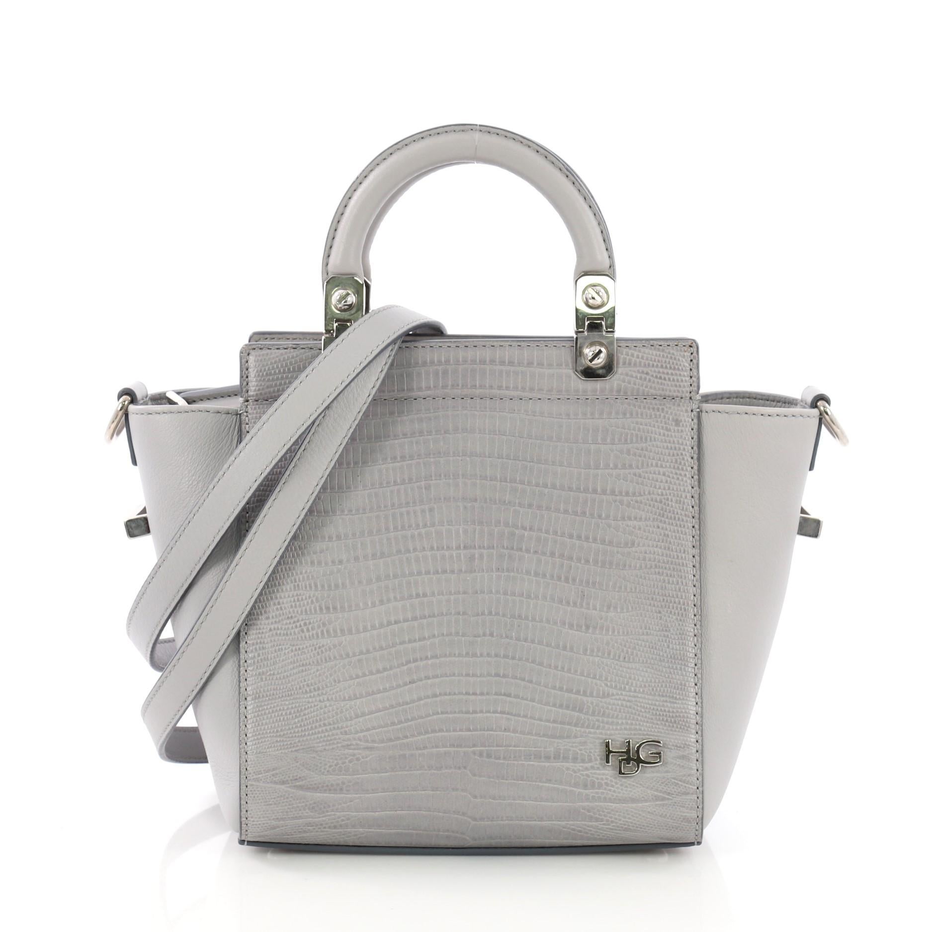 This Givenchy HDG Tote Lizard Embossed and Leather Mini, crafted in gray lizard embossed leather, features winged gussets, frame top, dual rigid leather top handles, a small HDG logo, and silver-tone hardware. Its zip closure opens to a black