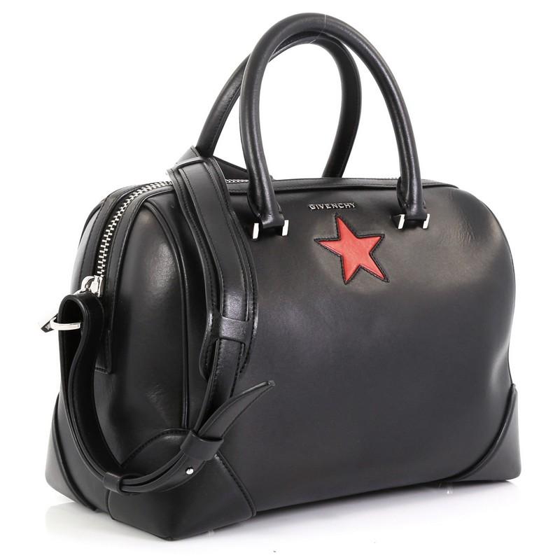 This Givenchy Lucrezia Duffle Bag Leather Medium, crafted from black leather, features a raised gold Givenchy logo, dual-rolled leather handles, a single red star across the front, and silver-tone hardware. Its zip closure opens to a black