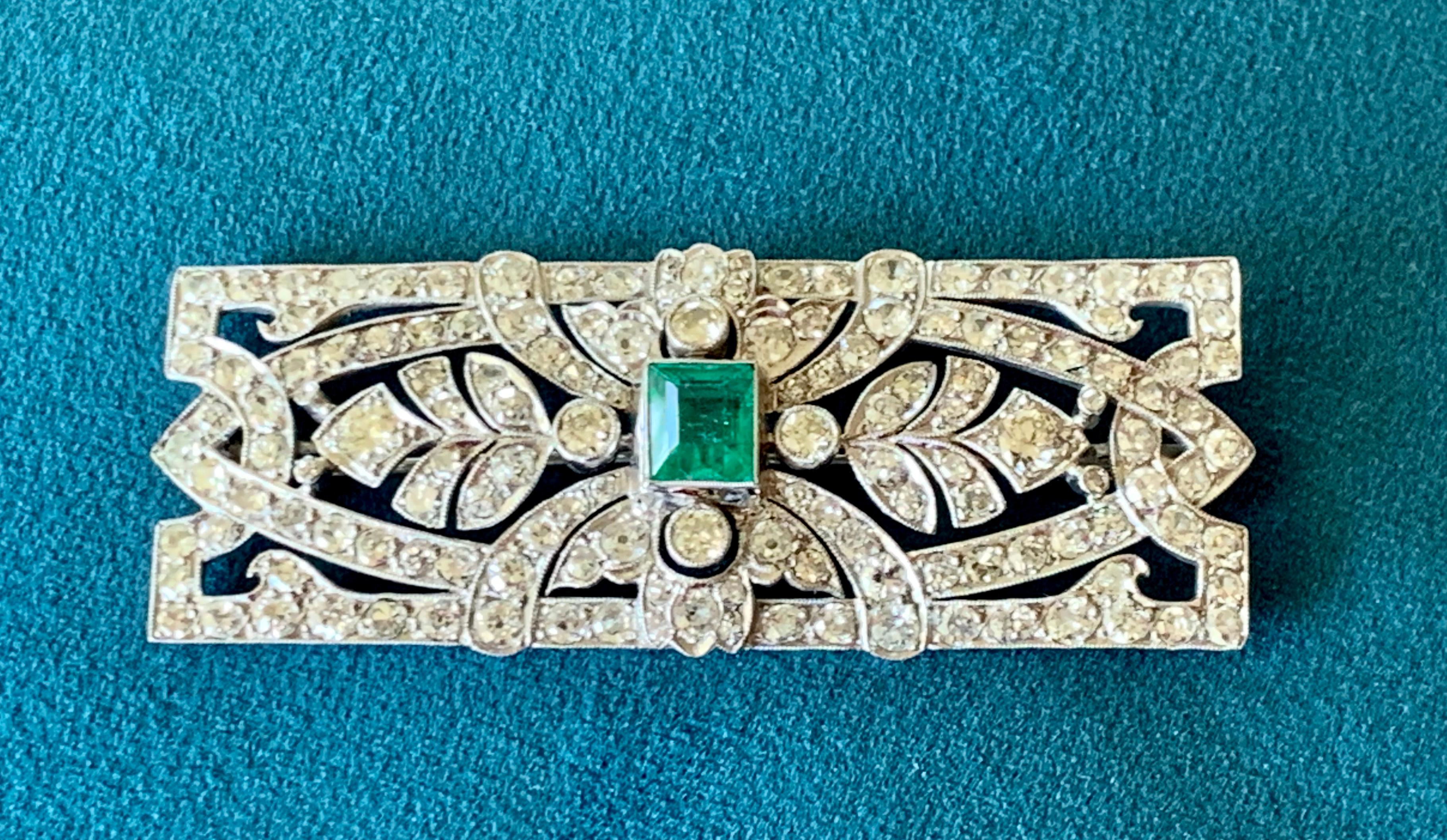 A splendorous original Art Deco brooch, circa 1920. A  vibrant green emerald of approximately 2 ct  is the center of attention of this quintessential Deco design finely crafted in platinum. The entire brooch is further embellished with many bright