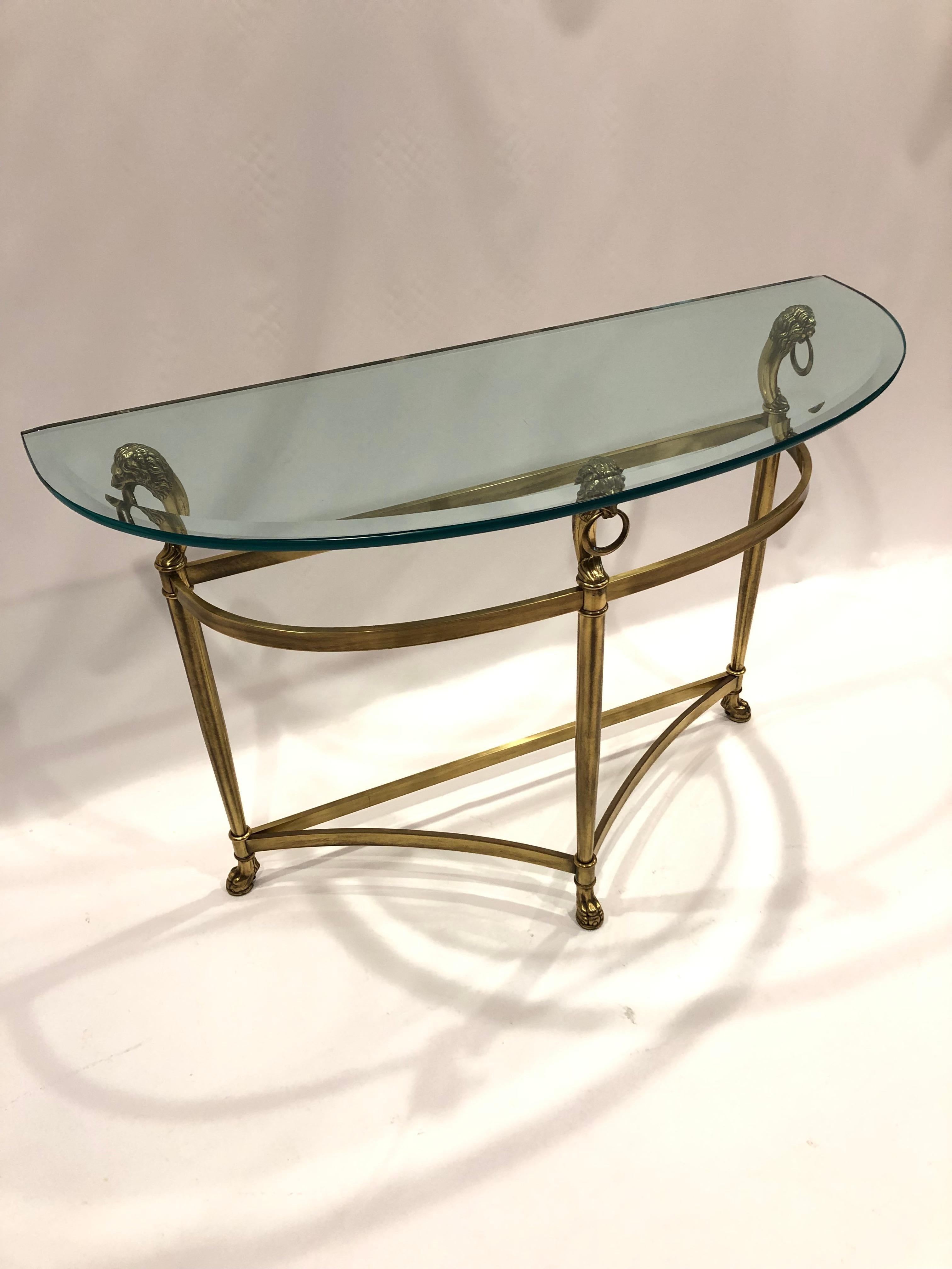 Stunning transitional demilune console table having brass base with handsome lion heads at the tops, paw feet and glass half moon bevelled top.