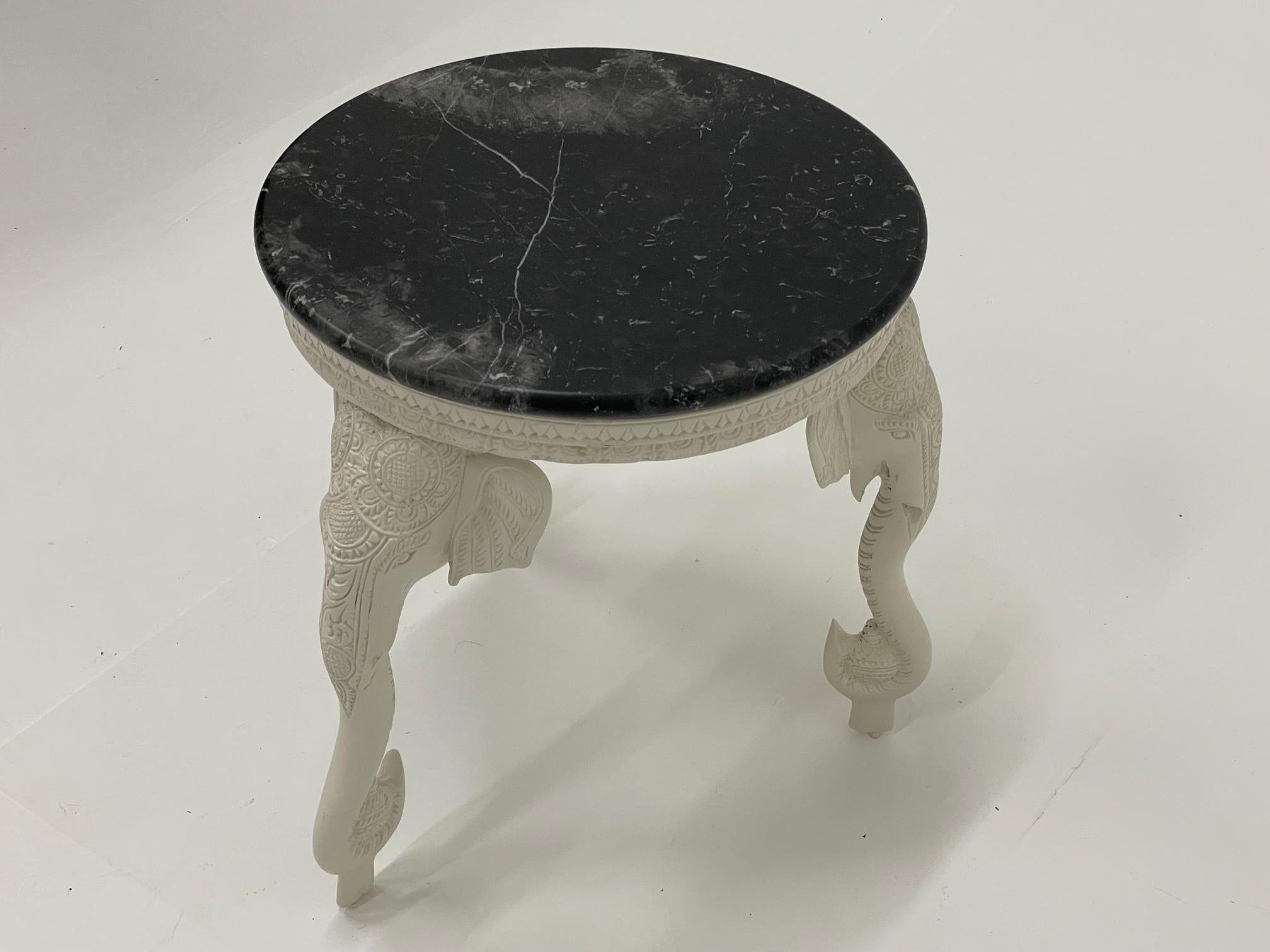 Chic round side table having meticulously detailed cream colored resin base with 3 elephant heads terminating in cleverly designed legs that are the elephants' curling trunks. A contrasting black marble top has some cream and grey veining.