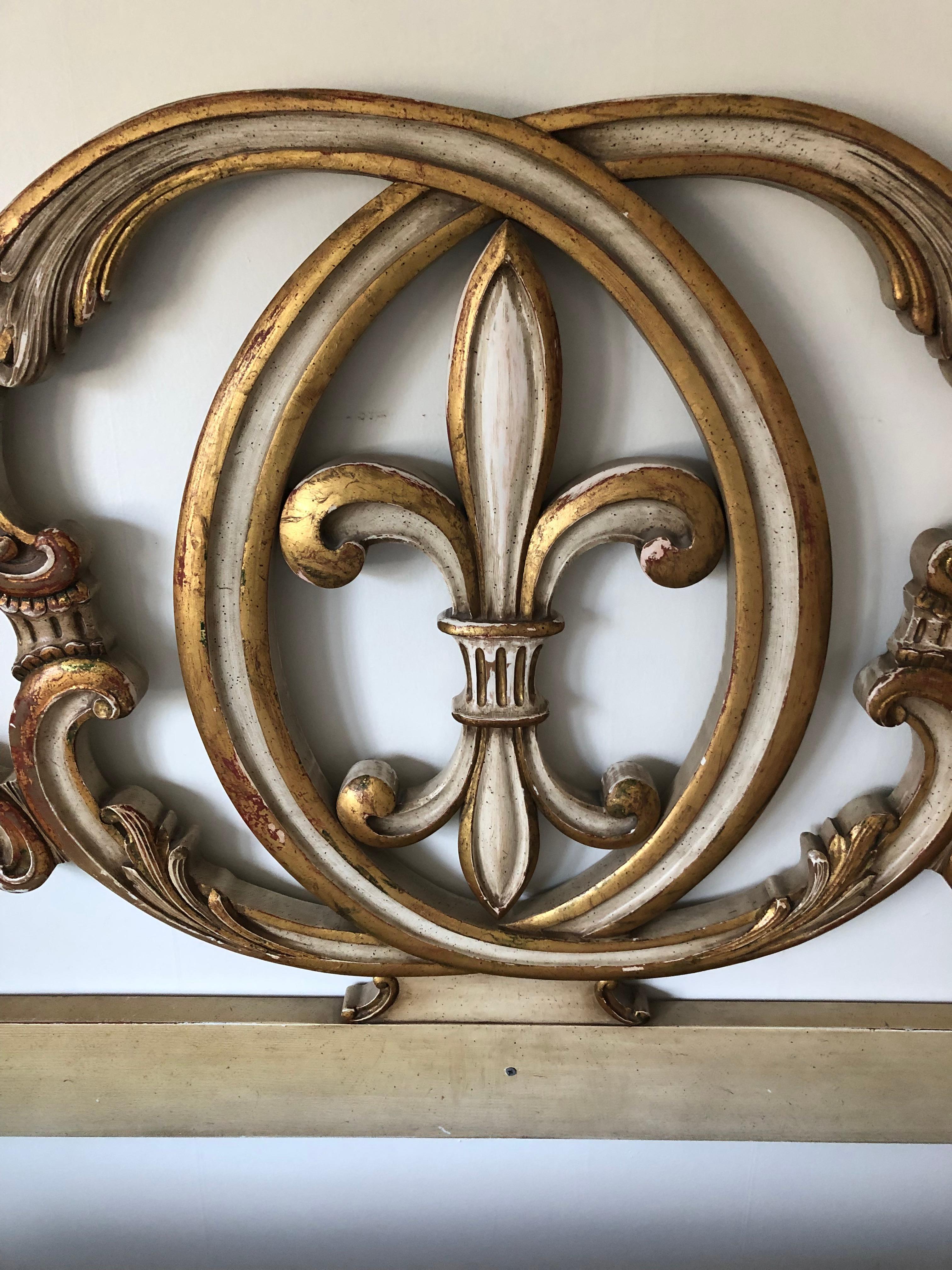 A rare find in a vintage glamorous king size headboard having carved and painted wood in an antiqued cream with gilding and some red underpaint visible. A central striking and timeless fleur di lis symbol sets the tone for this moviestar headboard.