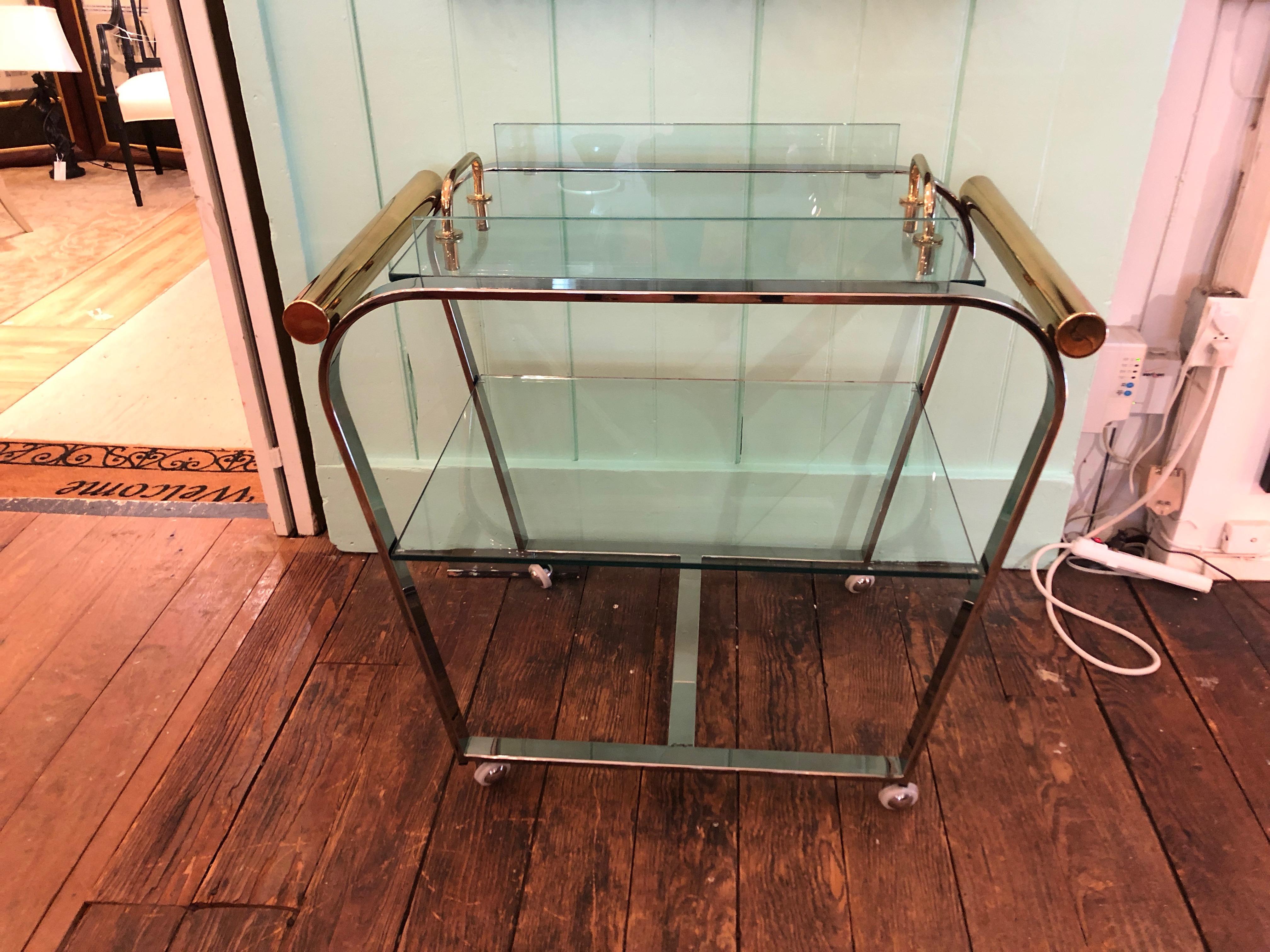 A gorgeous mixed metal midcentury bar cart with a rare deco-esque form, having two glass tiers with the top one being an ingenious heavy lift off tray with brass handles. On casters.
13 inches clearance between lower shelf and top
Tray is 25 W x