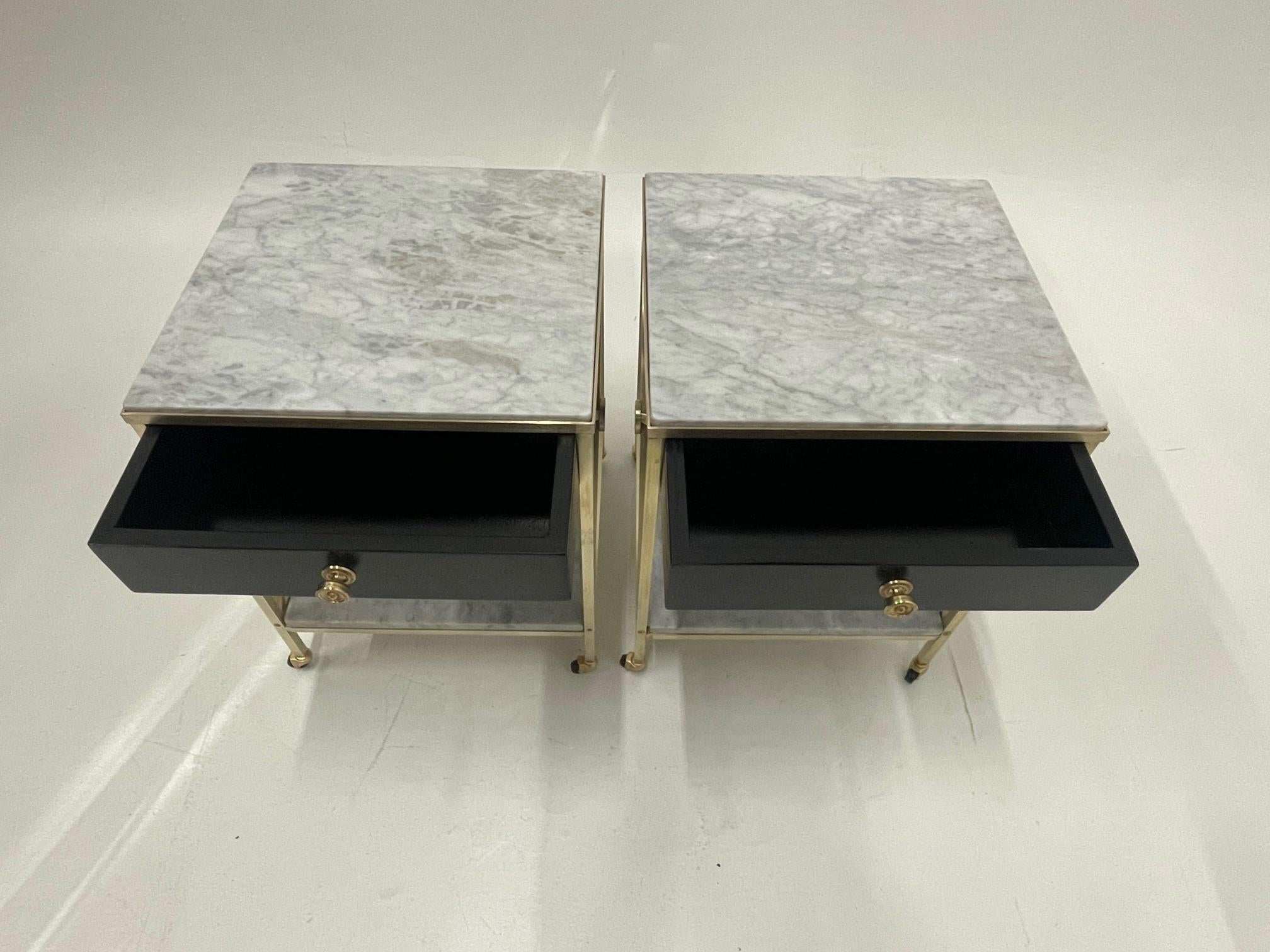 Sublime elegant pair of Maison Jansen style Hollywood Regency end tables or night stands having a striking combination of white marble and polished brass offset by ebonized wood black single drawers. Classy criss cross X designs grace the sides,