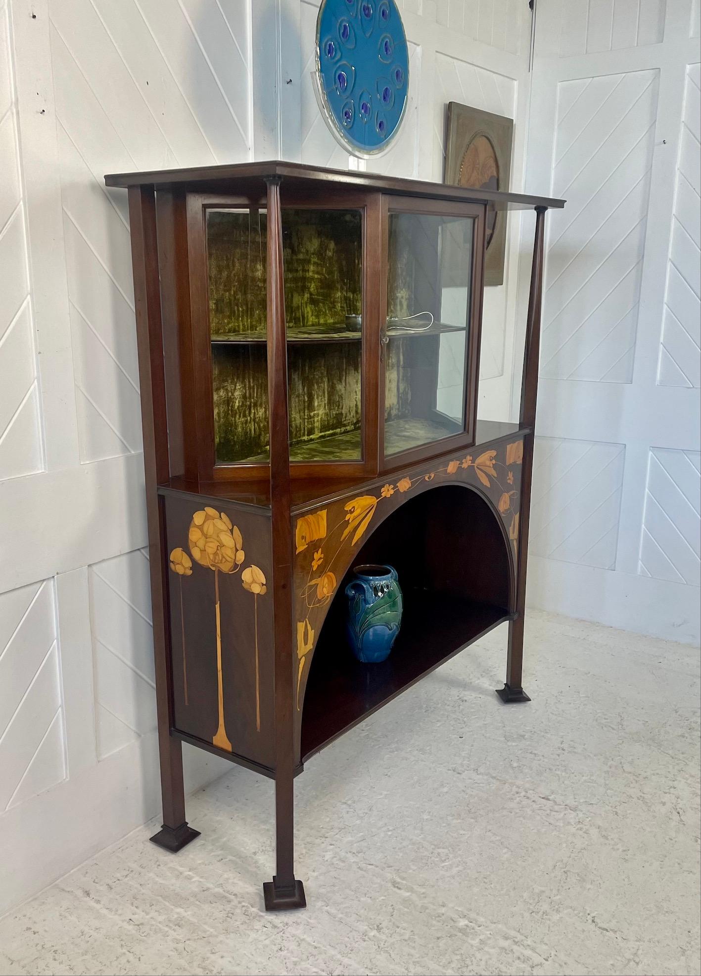 Glasgow School mahogany cabinet
With stylised trees, flower and foliage inlay design
Well proportioned canted glass display cabinet and original green crushed velvet interior
Voysey style uprights, flared feet and overhanging top
Lower display shelf
