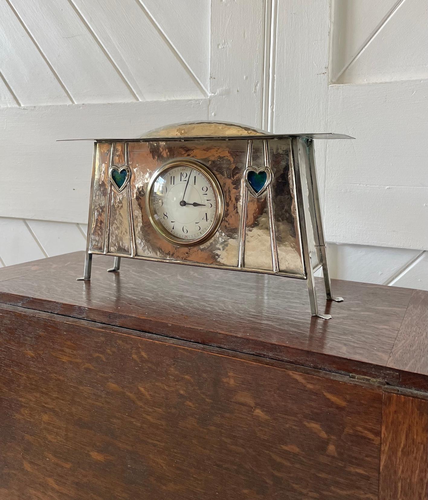 Glasgow School silver plate on copper mantel clock

Riveted with a domed top and set on 4 stylised splayed feet

Rare inset blue and green enamel heart decoration

Circular white enamel faced clock with a French Movement

Glasgow School

Circa 1900