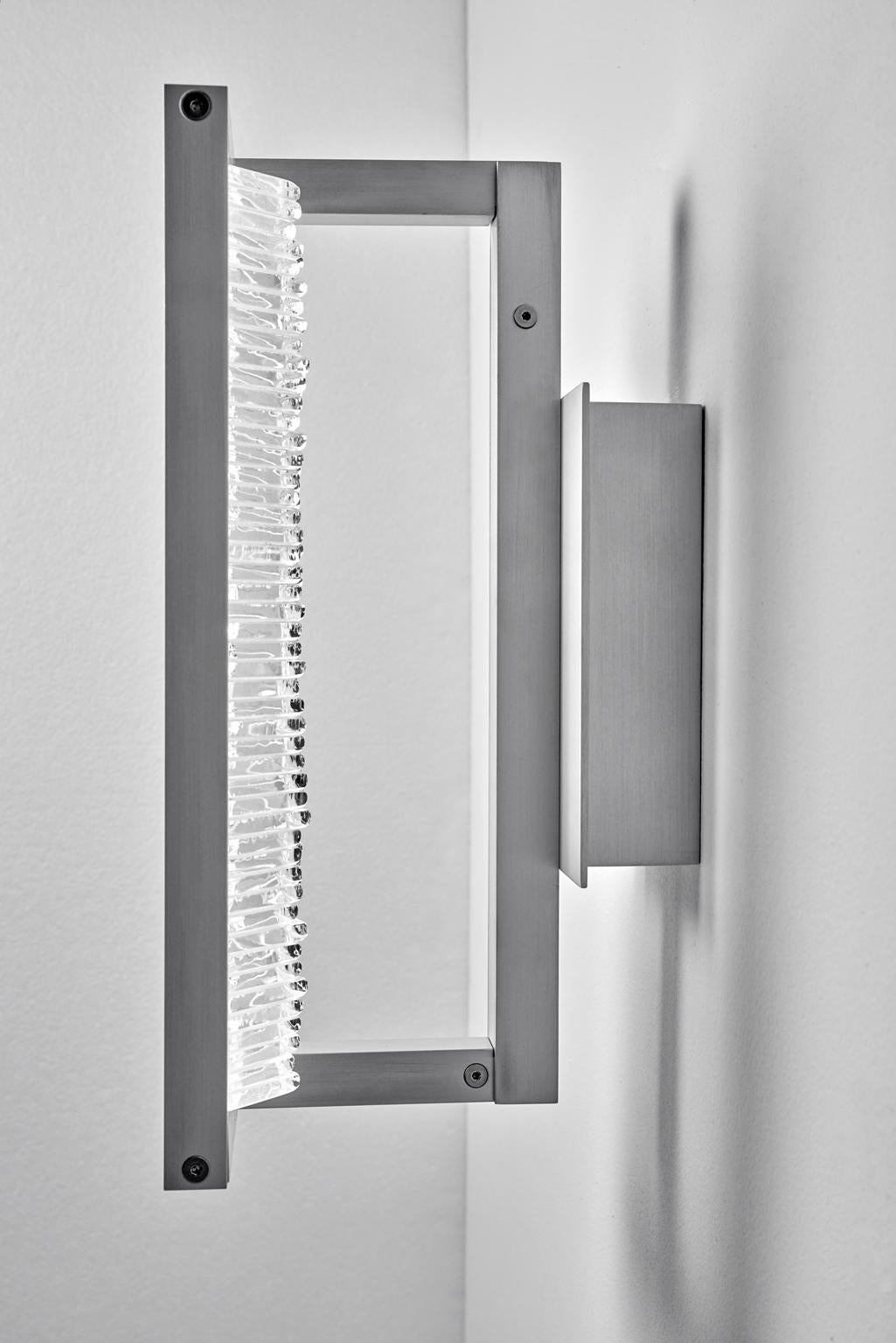 Designed and created by Sidney Hutter Glass & Light, this wall sconce has an ultra contemporary style that looks good in any environment. The sconce is framed with anodized aluminum. By laminating clear glass pieces together in a horizontal pattern