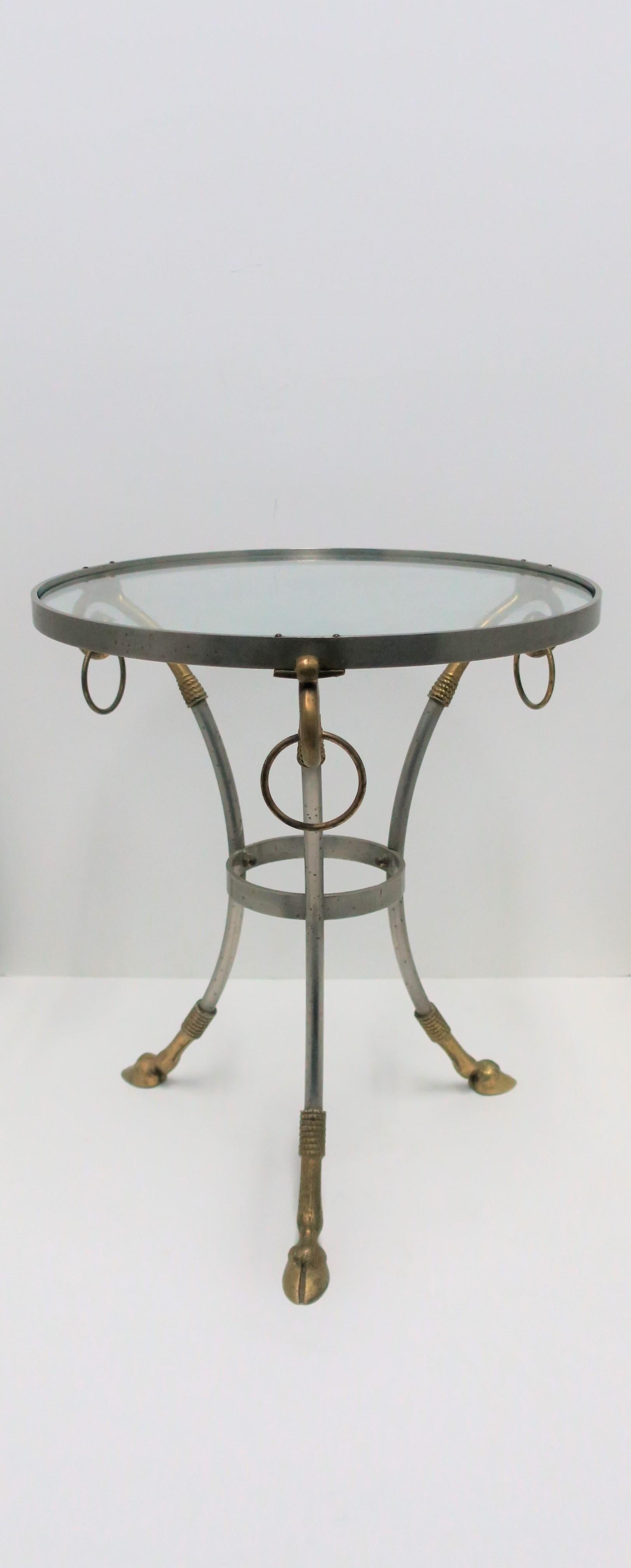 A small and substantial French Gueridon round side or drinks table with brass rams head hoof detailing, in the style of designer Maison Jansen, circa 20th century, France. Table is matte steel and brass with brass loop/ring detail at top, hoof/legs