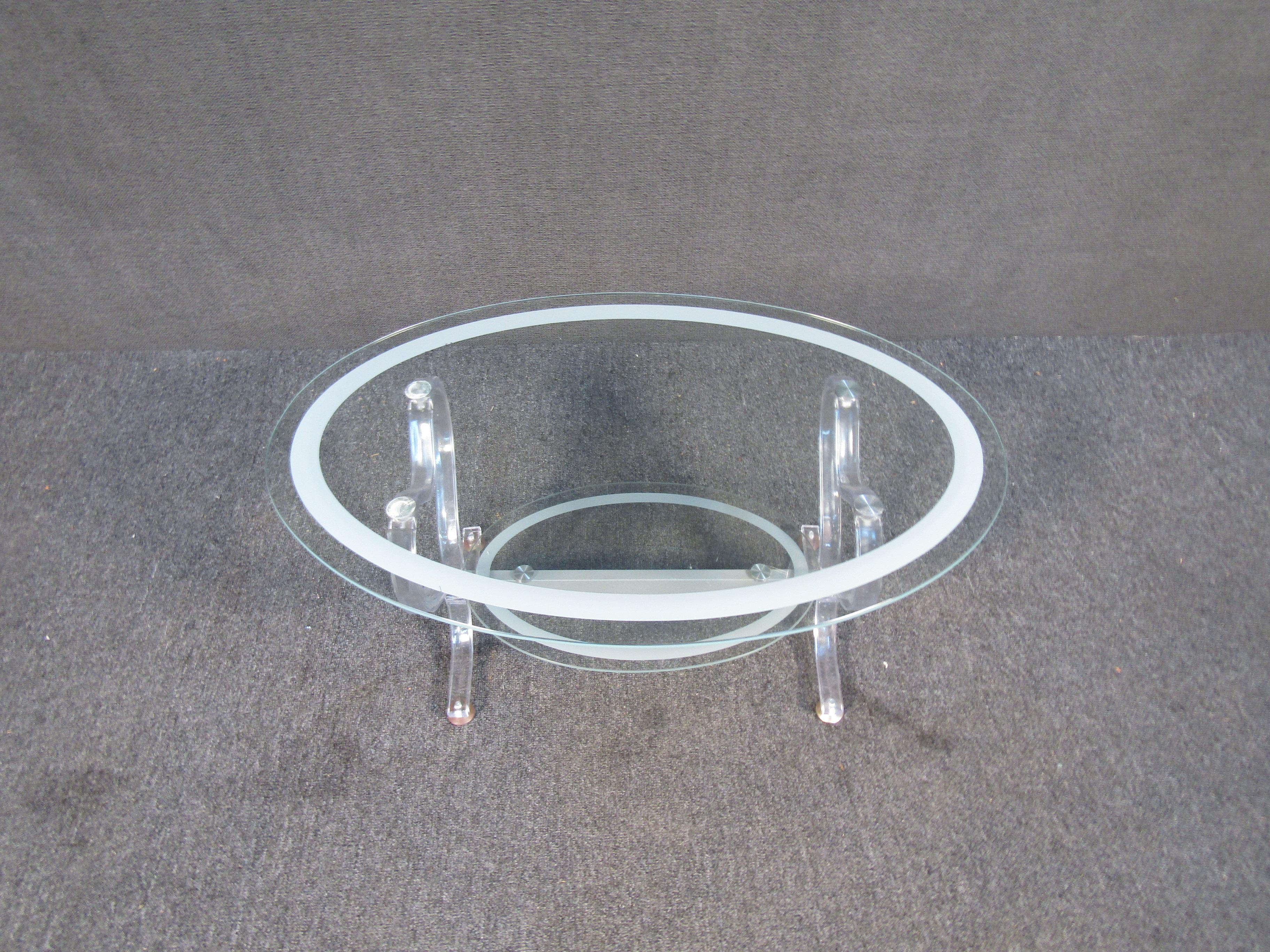 Cette table unique en verre à deux niveaux présente un design ovale et une base en Lucite ornée. Des accents de rayures blanches entourent les étagères du haut et du bas. Cette table est un ajout parfait à tout espace de détente nécessitant une