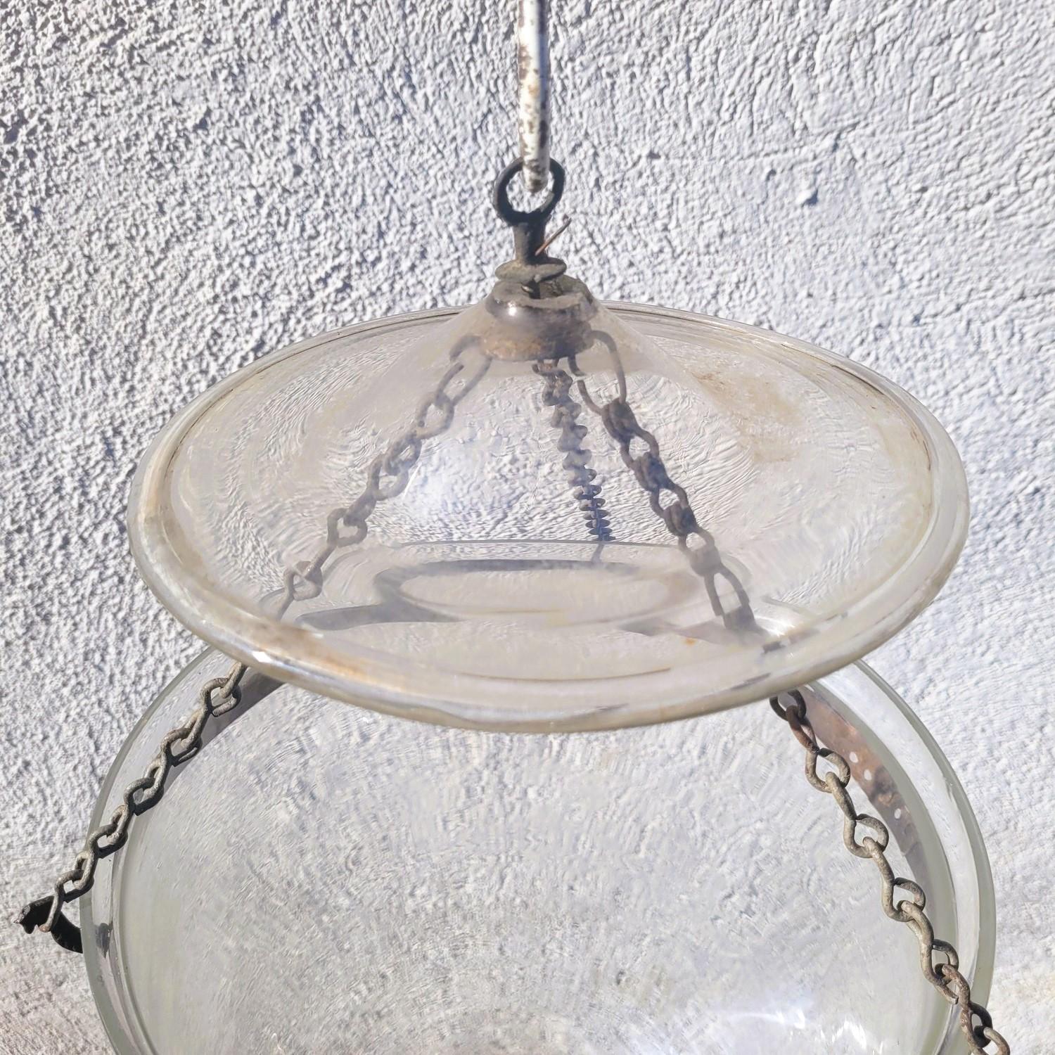 Lanterne en verre transparent en forme de cloche avec son récipient à fumée, chaînes et fixations en laiton gravé et têtes d'oiseaux
La cloche contenant une bougie ou une lampe à huile est maintenue par un cercle en laiton sur lequel sont fixés