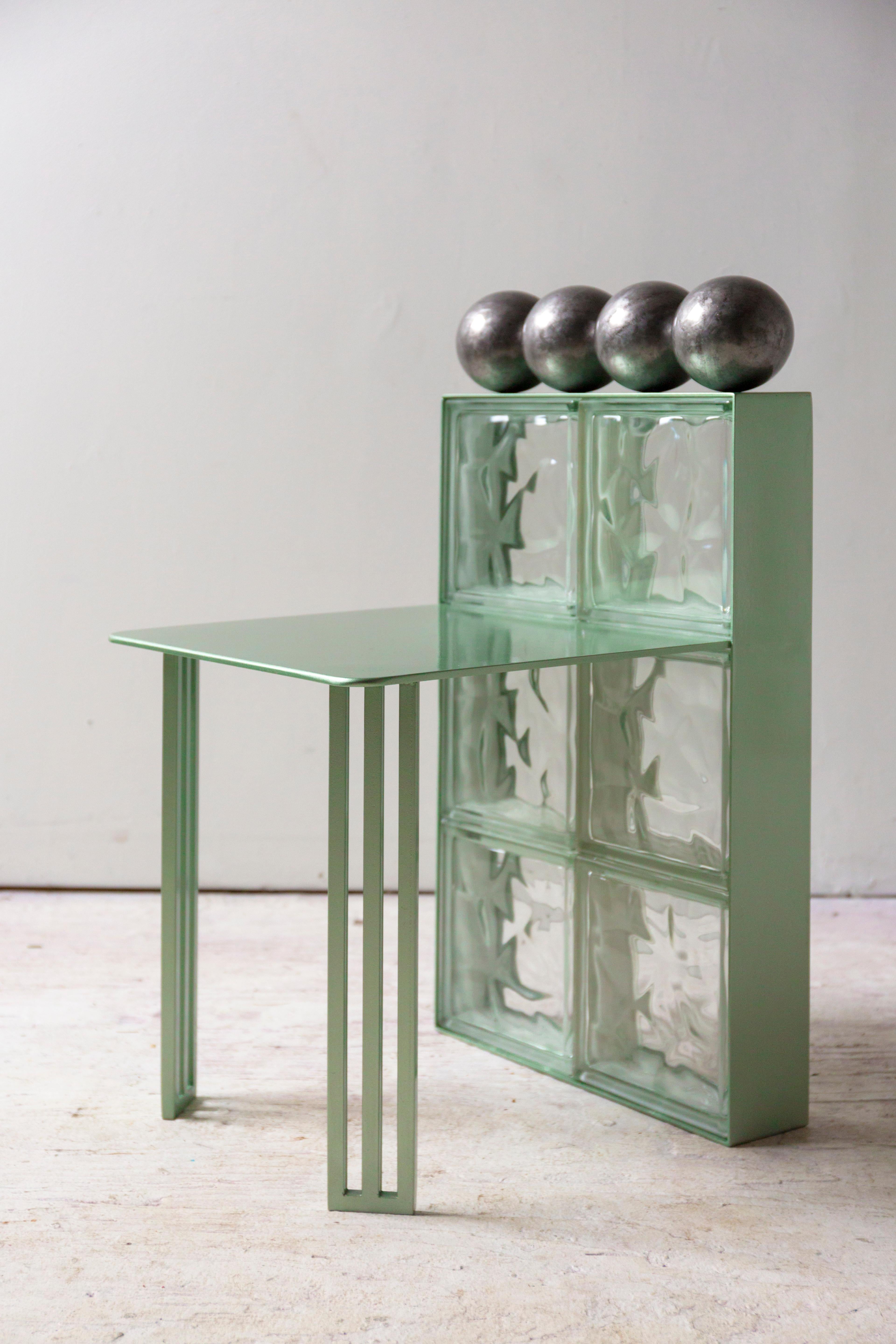 This compact steel chair is a nod to glass block architecture and playful geometry. Four welded iron balls create an illusion of balance along the solid glass block wall that acts as a back rest. The gentle green metallic finish highlights the