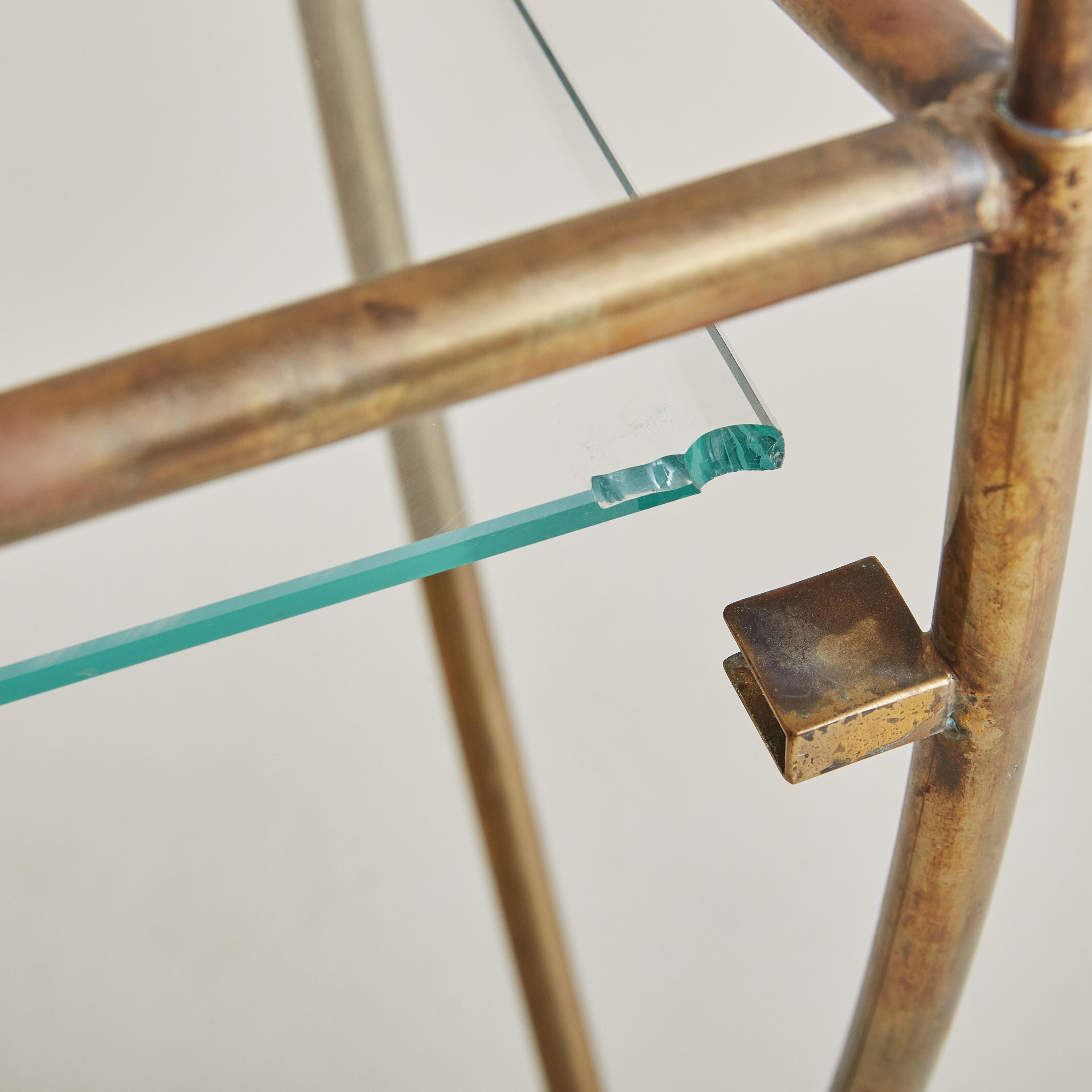 An Italian glass and brass side table reminiscent of Jean Royere’s ‘Yo Yo’ designs. This table features a delicate patinated brass frame with curved legs, a center stretcher, and an angled top adorned with finial details. The rectangular glass shelf