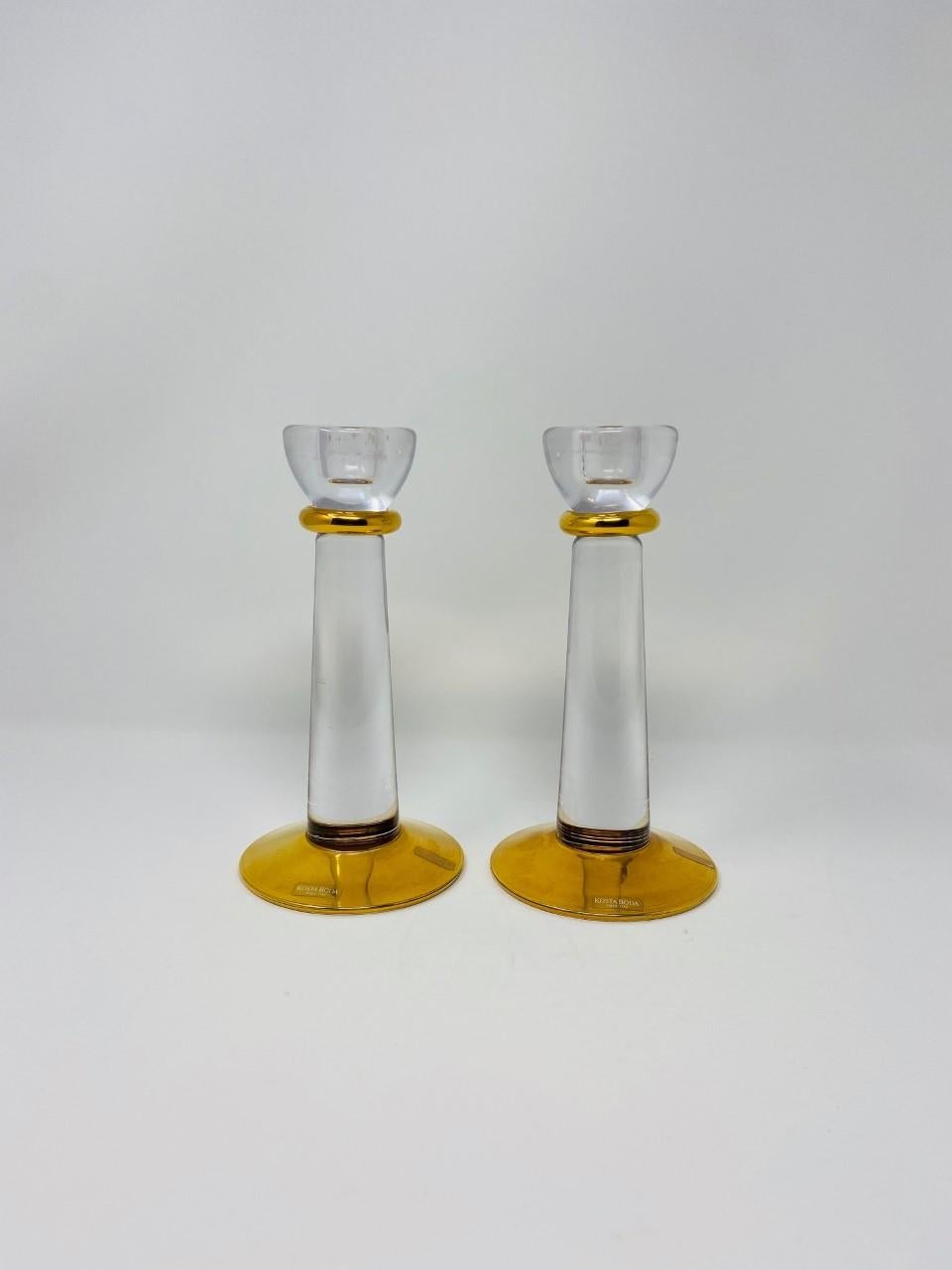 Minimalist and incredibly glamorous pair of Kosta Boda glass candle holders. The pair exhude beautiful lines that can streamline or maximize elegance in your décor. Crystal clear glass accentuated with a gold finish at the base and neck. Both pieces