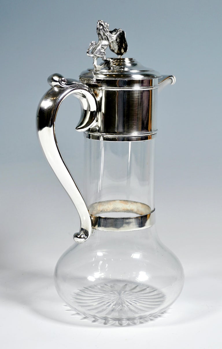 Carafe made of clear glass with a bulged stand and narrowed straight neck, cut bottom star, silver fitting over the upper area of the neck piece, volute-shaped handle, which is also attached at the lower end by a ring-shaped band around the carafe