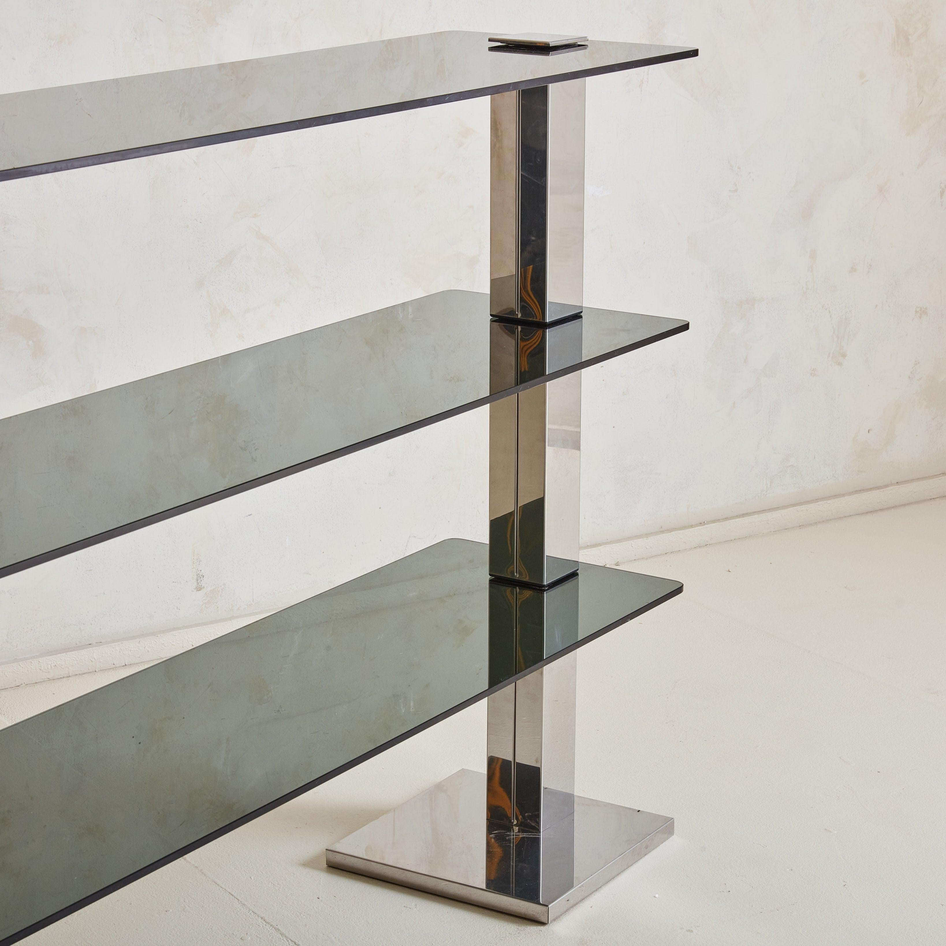 A 1970s Italian Modern console table or shelf featuring a chrome frame with two thick, rectangular legs and dramatic square feet. Three gray hued, smoked glass shelves with curved edges rest between the leg components. This piece requires placement